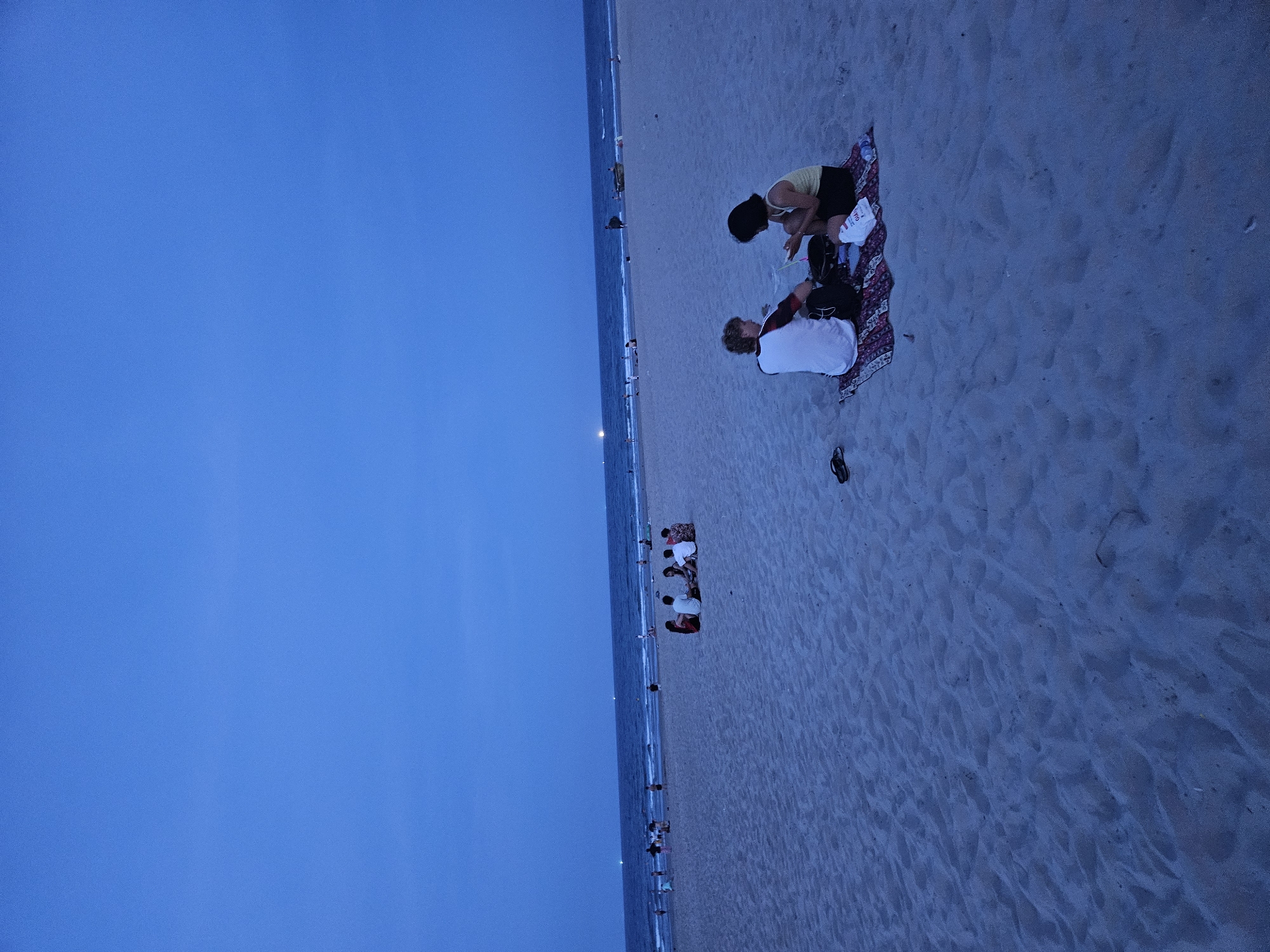 The image shows a wide, sandy beach in Đà Nẵng, Vietnam, at dusk. The sky is a deep blue, transitioning into a lighter blue at the horizon where the sea meets the sky. The sea is calm with gentle waves. In the foreground, two young people are sitting on a patterned mat on the sand, facing each other. They appear to be engaged in conversation or an activity together. The person on the left is wearing a white t-shirt with black and red accents, and the person on the right is wearing a yellow tank top and black shorts. There are some belongings near them, including a bag and a white plastic bag. Further down the beach, closer to the water, are several other groups of people, some sitting and some standing, indicating a public gathering. The sand is textured with footprints and natural undulations. The overall atmosphere is serene and peaceful, characteristic of a beach evening.