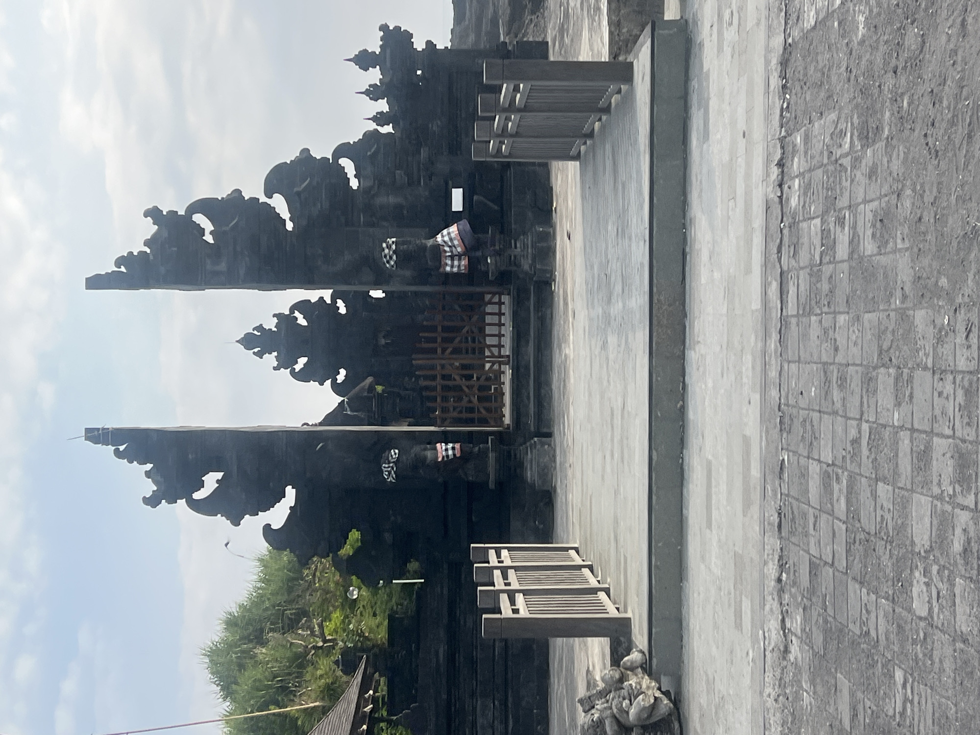 The image captures a view of the Taman Ayun Temple in Mengwi, Indonesia. In the foreground, a paved walkway with a checkered pattern leads to a raised platform, bordered by wooden railings on either side. The platform is made of large, grey rectangular tiles and has a concrete base. To the left of the walkway, a large, carved stone statue is visible.
Beyond the platform, a grand, dark stone gateway dominates the scene. This gateway features intricate carvings and split architectural elements, resembling wings or ornate spires. Two statues, adorned with traditional Balinese checkered cloths, stand on either side of the gateway's entrance. The entrance itself is partially blocked by a wooden gate. Behind the gateway, more dark stone structures and rooftop details of buildings are visible, interspersed with lush green foliage on a hill. The sky above is overcast with light clouds. The overall impression is of a significant cultural and religious site.