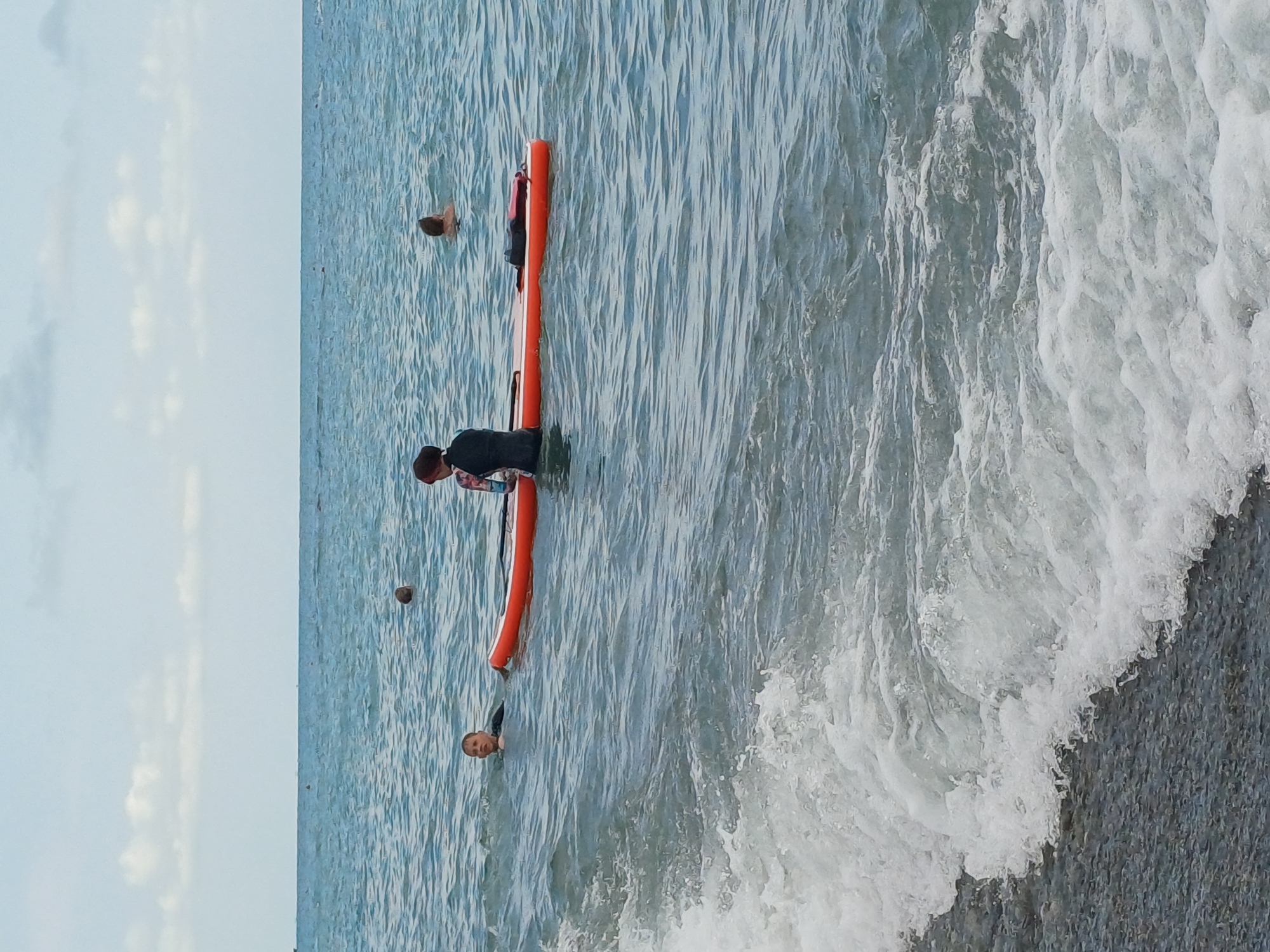 A group of people are in the Black Sea near the shore in Sochi, Russia. In the foreground, waves crash onto a pebble beach. Further out, a bright orange paddleboard is in the water. Two individuals are near the paddleboard, one appears to be sitting on it while wearing a black wetsuit, and another person is in the water next to the board. Two other individuals are visible in the water further offshore, with only their heads showing. The sky is overcast with scattered clouds.