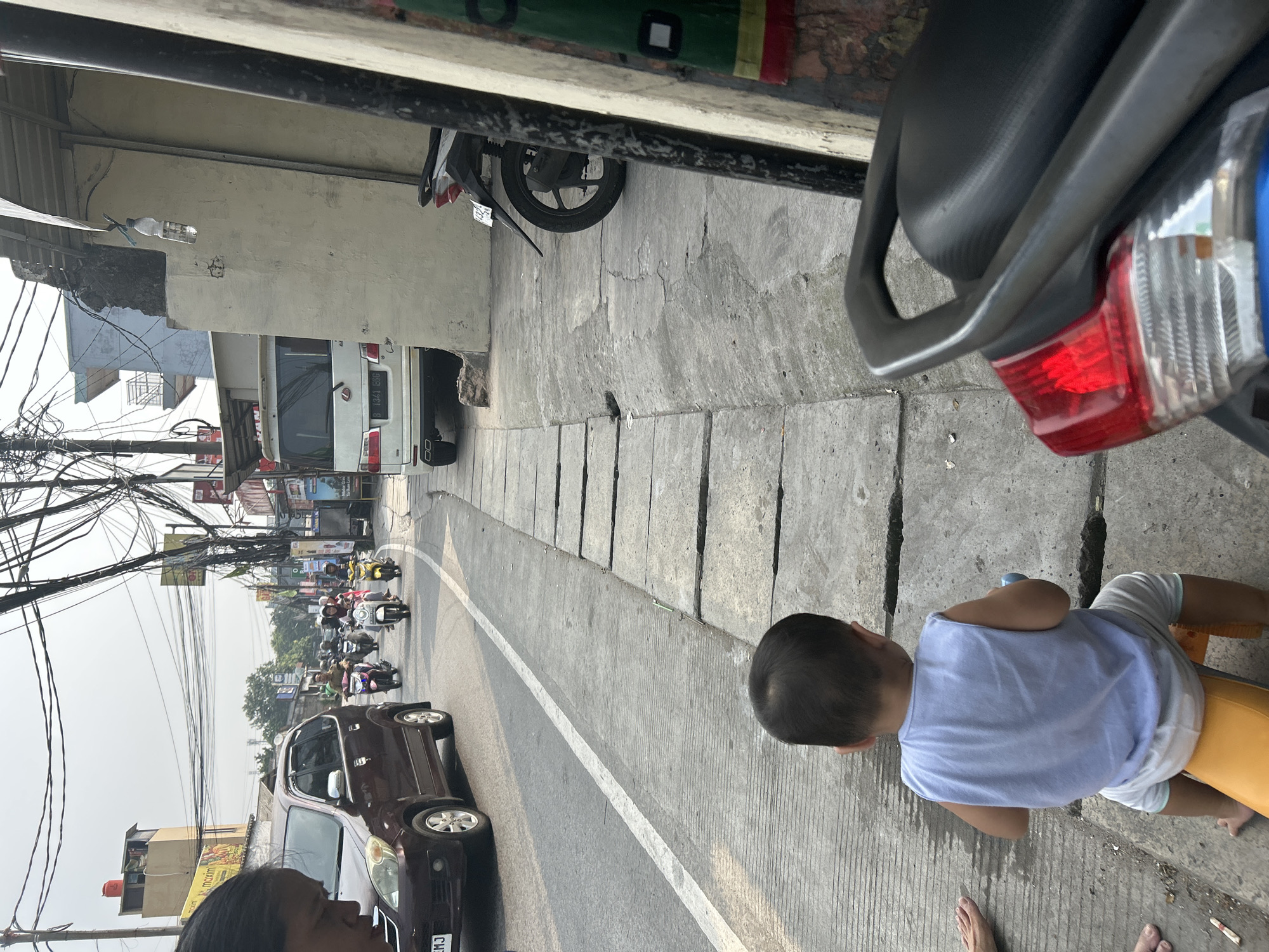 A low-angle, outdoor, eye-level shot captures a street scene in Depok, Indonesia, on a bright, overcast day. In the foreground, from the back, a young child wearing a light blue tank top and white shorts is seated on a yellow toy vehicle. The child's head and upper back are visible. To the left of the child, a person's bare feet and toes are visible, suggesting they are barefoot and standing or sitting on the sidewalk. A dark-colored motorcycle is parked on the right side of the frame, with its tail light illuminated and a part of its black seat in view.
The scene depicts a busy urban street. A dark red minivan is driving in the left lane of the road, with its front end partially visible. Several motorcycles, some with riders wearing helmets, are moving along the road in the distance. The road is paved with asphalt and has a white lane marker. A concrete sidewalk with square slabs runs along the right side of the road, adjacent to a building. The building has a neutral-colored wall with some visible damage and wires hanging from it. Overhead, a dense network of electrical wires crisscrosses the sky. In the background, there are more buildings, trees, and street signs, indicating a commercial or mixed-use urban area. The overall atmosphere suggests a typical day on a moderately trafficked street.