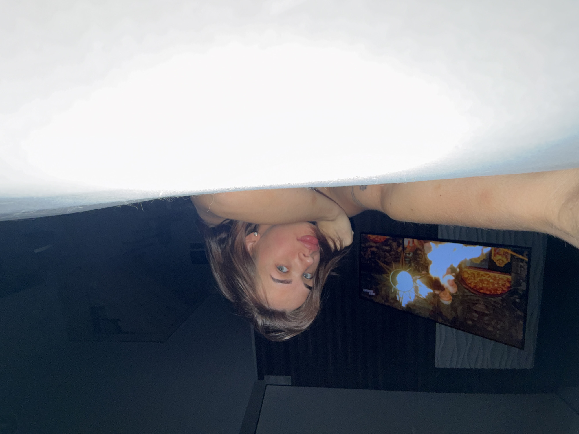 The image depicts a person, likely female, upside down, partially obscured by what appears to be the underside of a light-colored surface, possibly a bed or table. The person is facing the camera, with hands positioned near their head. In the background, a television screen displays an image that appears to be a scene with fire and food. The environment seems to be an interior space, likely a room, with dark walls. The location in the city of Lobanovo, Russia, is not evident within the image content.