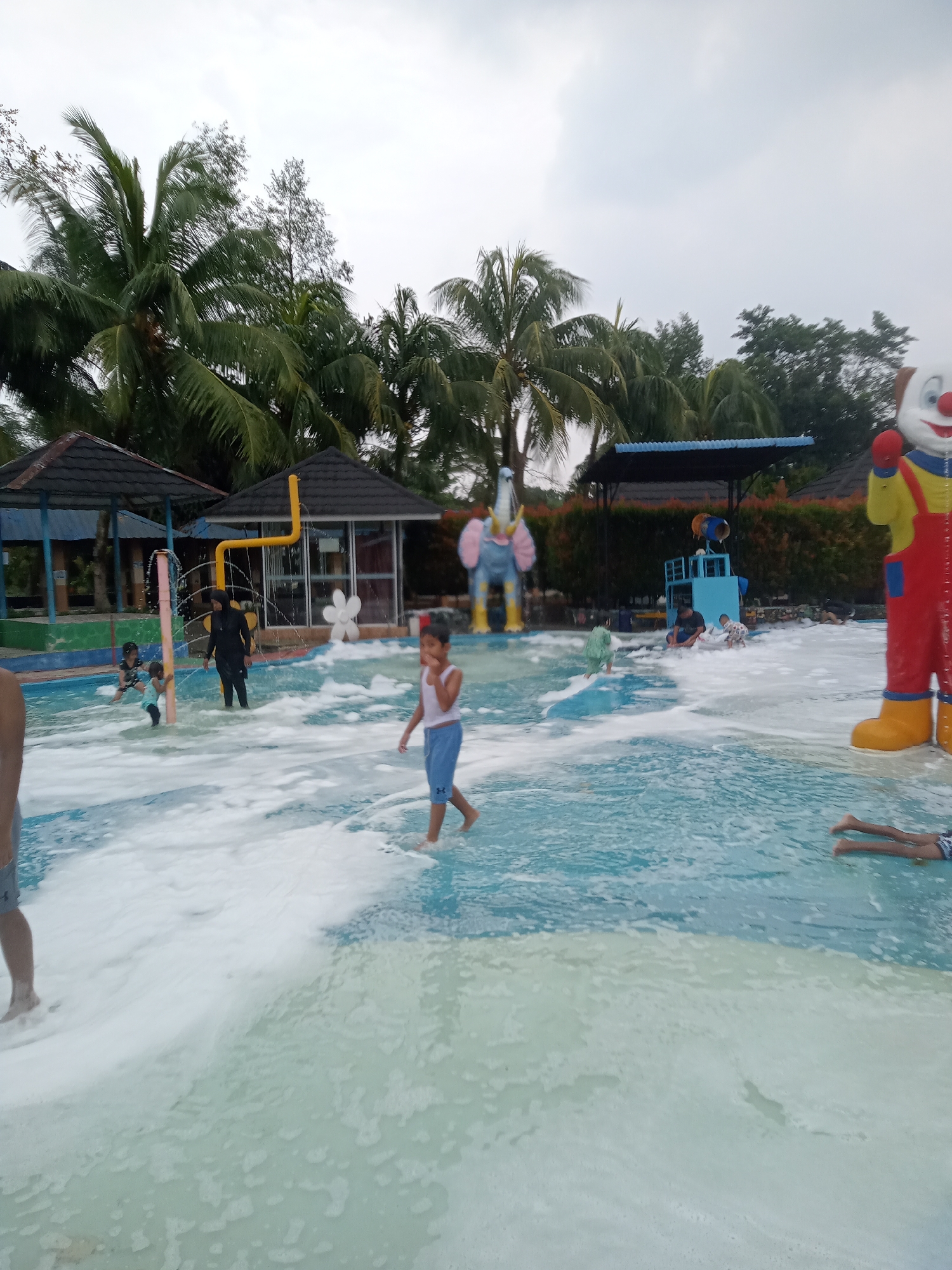 The image depicts a water park in the city of Baru, Indonesia. In the foreground, a young boy in blue shorts and a white tank top walks through shallow water filled with foam. To the left, the legs of another person are visible in the water. The middle ground shows several children and an adult in the water, some playing near a pink and yellow water feature. Further back, there are small covered seating areas with dark roofs and tiled walls. Behind these structures, tropical palm trees and other foliage are abundant, suggesting a warm climate. In the background, a large, colorful clown statue stands prominently on the right, and a blue elephant-shaped structure is visible on the left. The sky is overcast, indicating a cloudy day. The overall scene is one of a recreational water park with a lively atmosphere, featuring various attractions and people enjoying themselves.