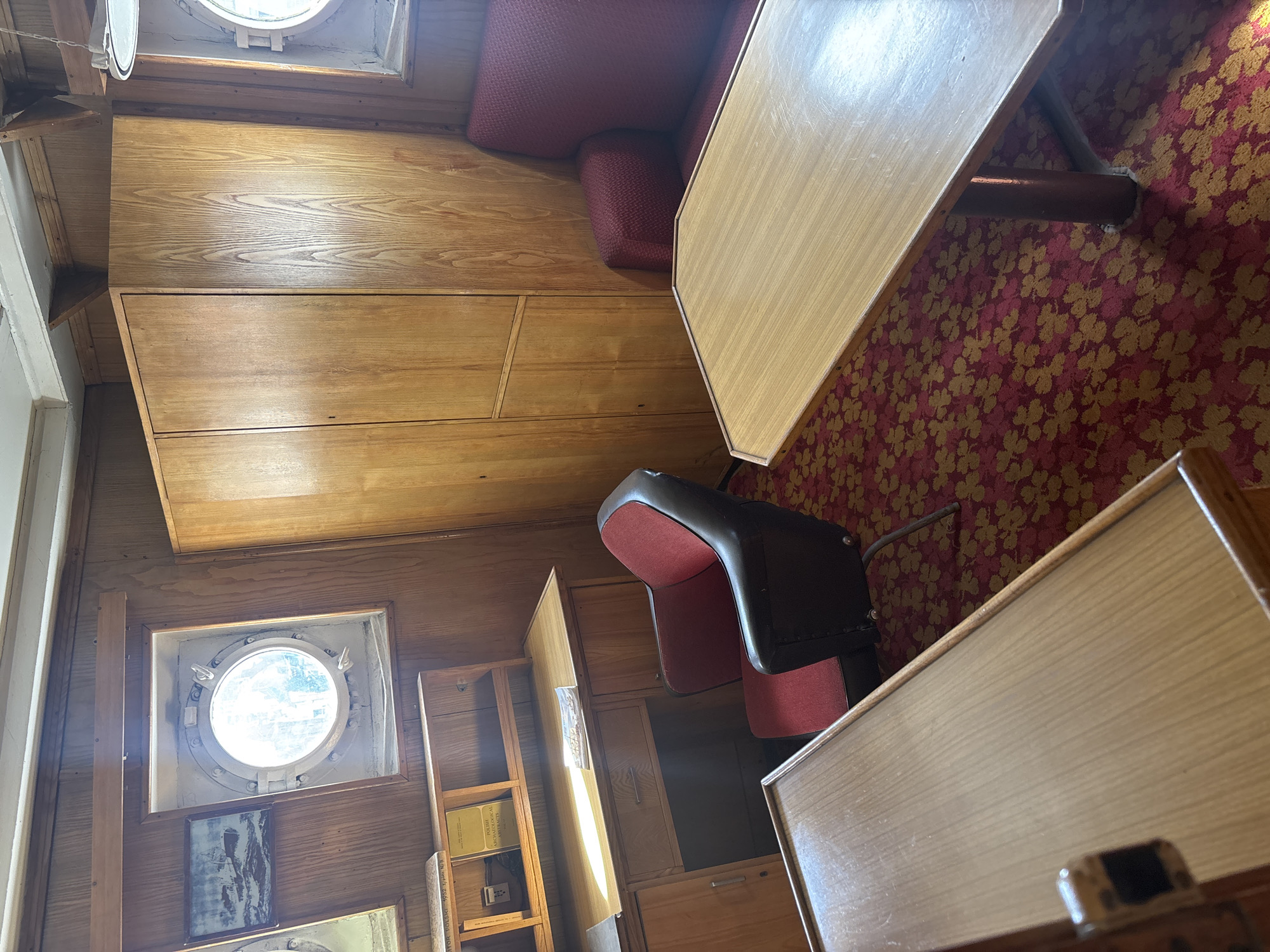 The image depicts the interior of a room with wooden paneling, a red patterned carpet, and a large, light-colored table. The room appears to be a dining or lounge area, possibly on a boat or in a vintage building.
On the left side of the image, there is a porthole window with a circular frame. Below the porthole, there is a framed picture. Further down, another framed item is visible. A wooden shelf unit is also present, containing books and what appears to be a small electronic device.
In the center of the image, a dark chair with red upholstery is positioned. Beyond the chair, there is another large table, this one with a light wood finish, similar to the table in the foreground. A red upholstered bench or sofa is partially visible behind the first table.
The lighting suggests it is daytime, with natural light streaming in from the porthole and possibly another window out of frame. The overall impression is one of a retro or vintage aesthetic. No people are visible in the image. The scene is still and quiet, with no discernable activity. Visible text includes titles on some of the books, such as "POLISH ARCHAEOLOGICAL" and "POLISH ARCHEOLOGICAL."