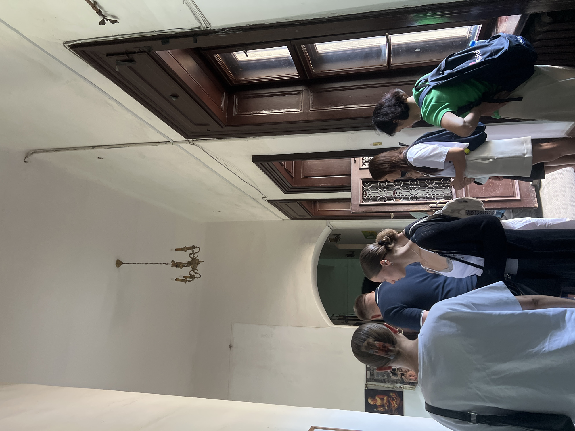 In an interior hallway, likely in the city of Karpaty, Ukraine, a group of five individuals, appearing to be tourists or students, are standing and walking. The hallway features high ceilings, white walls, and dark wooden trim around doorways and windows. A vintage chandelier hangs from the ceiling. To the right, large, dark-paned windows overlook an exterior. To the left, framed artwork is visible on the wall. The people are dressed in casual attire, with some carrying backpacks. One woman in the foreground faces away from the camera, wearing a white t-shirt and a dark shoulder bag. Next to her, a woman in a white tank top and black cardigan is also seen from the side. Further down the hallway, a man in a blue shirt stands with his back to the camera. Two other women are also present, one wearing a white t-shirt and light-colored skirt, and another in a green t-shirt and light-colored pants, both with backpacks. The scene suggests a moment of transition or observation within a historic or cultural building.