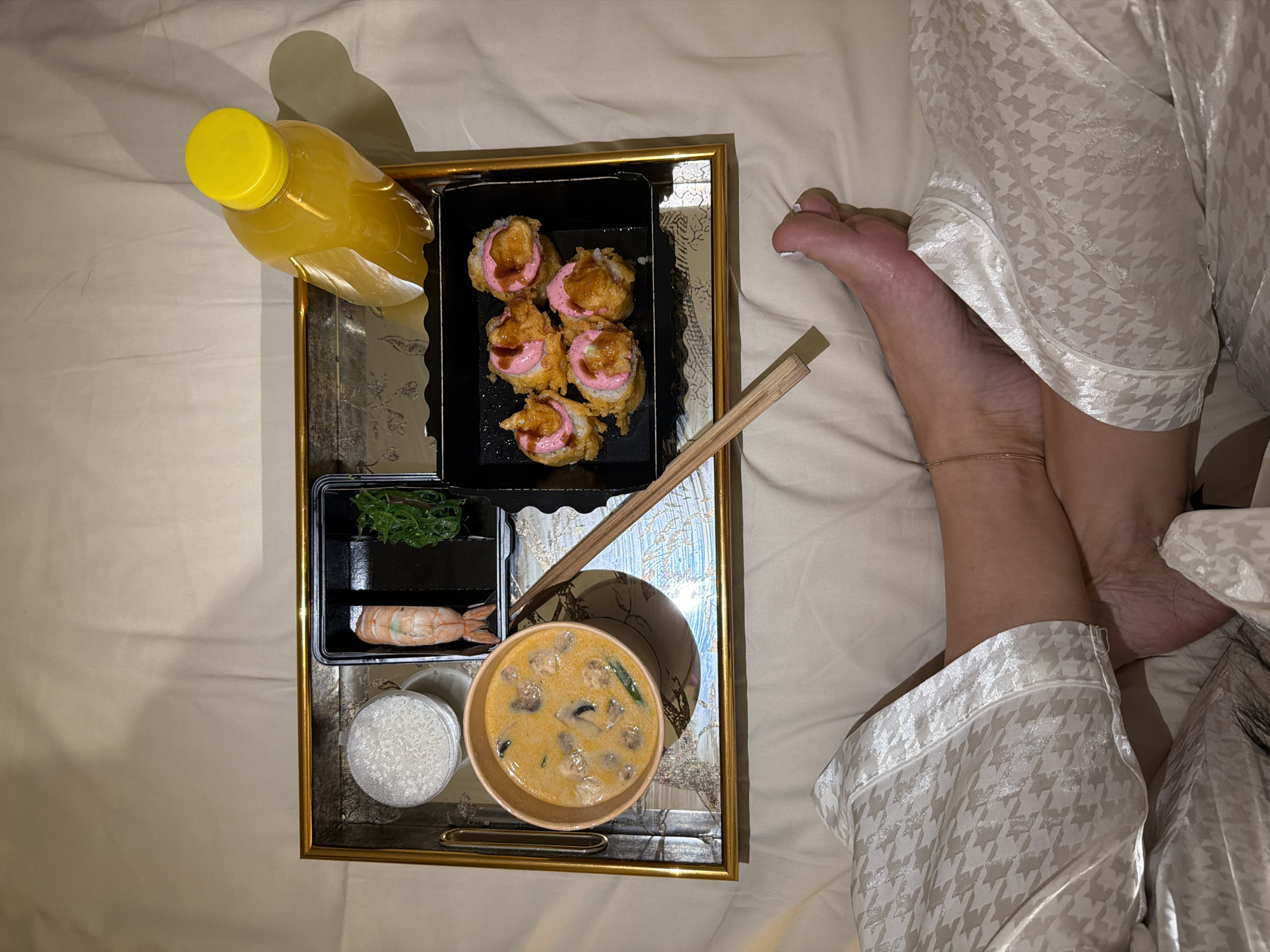 In Moscow, Russia, an overhead view captures a person's lower body and a meal service tray positioned on a bed. The individual's legs and feet are visible, partially covered by light-colored loungewear featuring a houndstooth pattern. An anklet adorns the right ankle, and toenails are painted white.
A rectangular serving tray with a golden frame and a patterned, reflective surface rests on the light-colored, wrinkled bed sheets. On the tray, a meal consists of a clear plastic bottle with a yellow cap, containing an amber liquid. A round, clear plastic container holds white rice. A black compartmentalized container holds a single cooked shrimp and a portion of green seaweed salad. A light orange bowl contains a creamy soup with mushrooms and green herbs. Another black container holds five pieces of what appear to be deep-fried sushi rolls, each topped with a pink sauce and a brown glaze. A pair of wooden chopsticks rests diagonally on the tray. The scene depicts a prepared meal for consumption in bed.