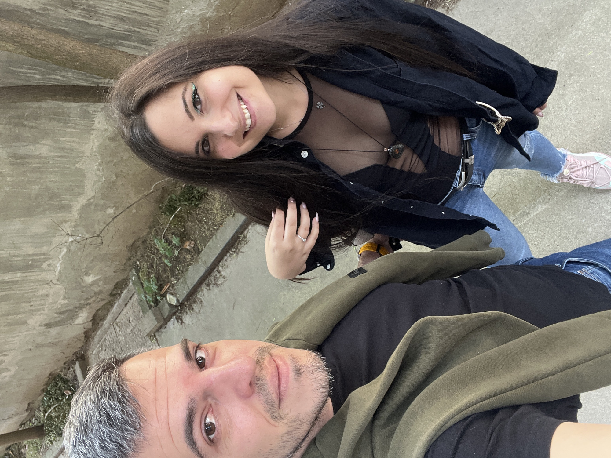 In an overhead selfie taken outdoors, a man and a woman pose for the camera. The man, on the left, has grey hair and a beard and wears a green sweater over a black t-shirt. The woman, on the right, has long dark hair, makeup, and a black sheer top with a black jacket. She is wearing blue jeans and pink sneakers. The background shows a concrete wall, a tree, and some greenery. The scene appears to be on a street in Sofia, Bulgaria, possibly near a park or a less maintained area of the city.