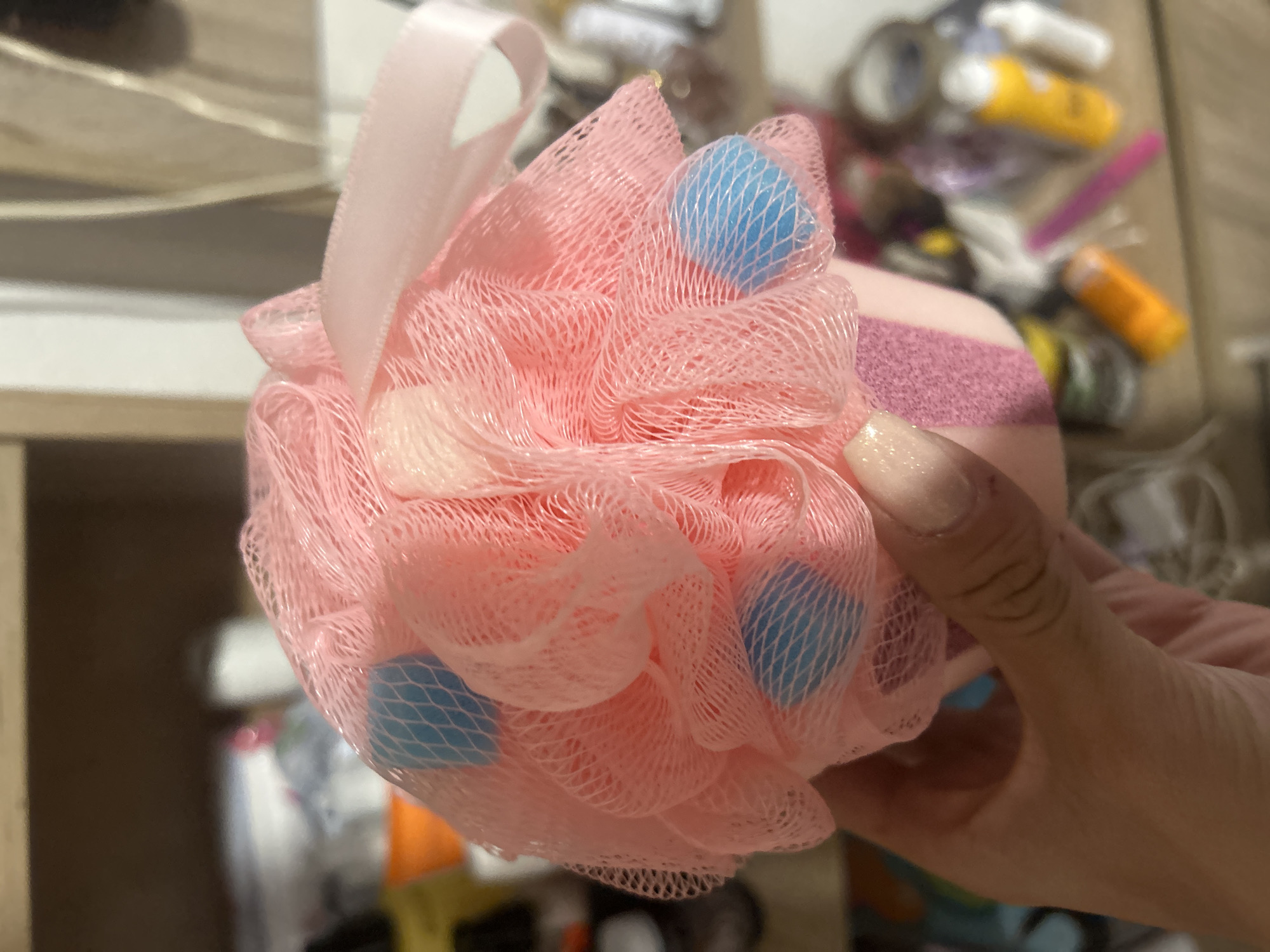 The image shows a close-up of a person's hand holding a pink mesh loofah with blue accents. The loofah is fluffy and textured, with a thin pink ribbon attached to the top. The background is blurred but appears to be a shelf with various toiletries and personal care items, including bottles and tubes. The lighting is warm and appears to be indoors, possibly in a bathroom or on a vanity. There is no discernible indication of the city of Perushtitsa, Bulgaria within the image itself.