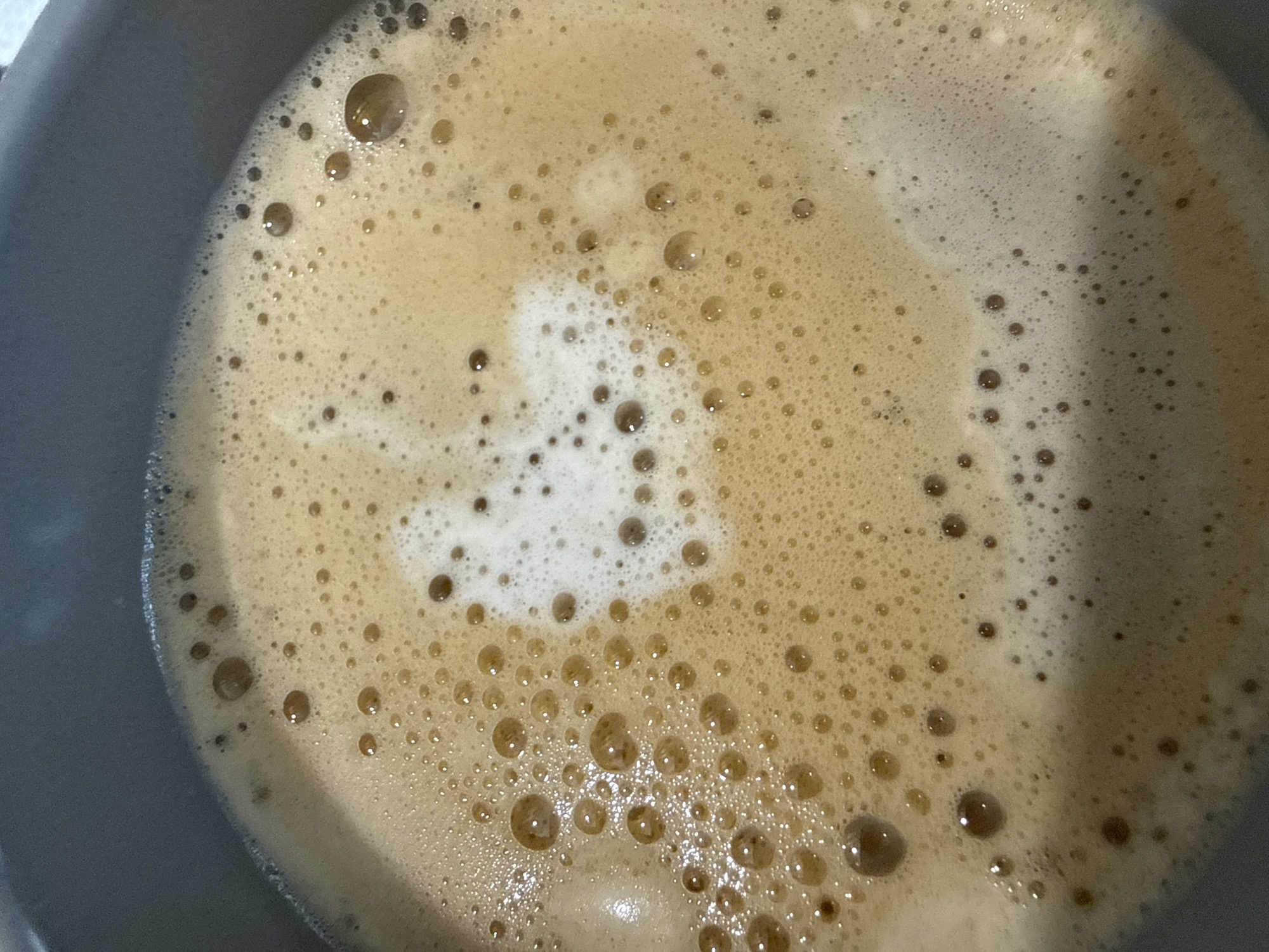 The image is a close-up, overhead view of the foamy surface of a beverage, likely coffee, in a dark gray mug or pot. The foam is a light tan color with a concentration of smaller bubbles that gradually transition to larger bubbles, particularly towards the left and bottom of the frame. A distinct lighter area of froth, forming a roughly heart-shaped pattern, is visible in the center. The beverage surface is uneven, with varying textures and depths of foam. The dark gray rim of the container is visible along the top and left edges of the image, indicating the close proximity of the camera to the surface of the liquid. There is no visible indication of people, actions, or interactions. The location in the city of Varna, Bulgaria, is not discernible from the image itself, as it contains no environmental or identifying landmarks.