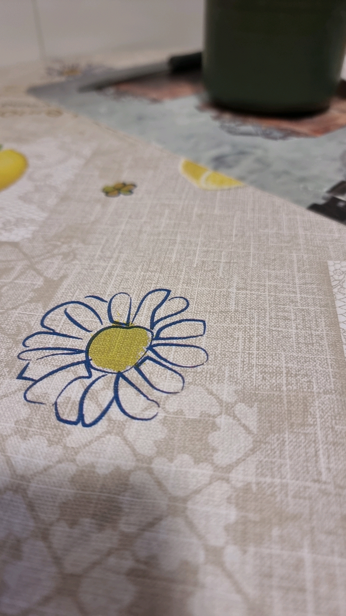 A close-up shot of a tablecloth features a stylized blue daisy with a yellow center. The tablecloth has a light beige background with a subtle grid pattern and faint floral motifs. To the upper right, a dark green object, possibly a cup or container, is partially visible. The scene is indoors, with shallow depth of field.