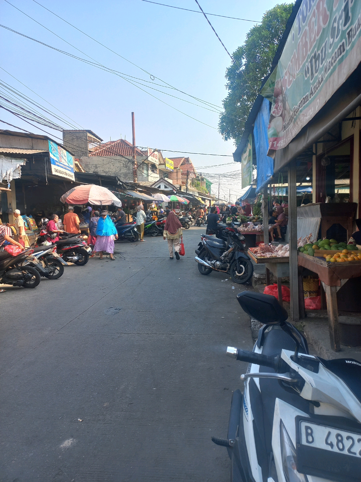 This is a street-level, medium-angle photograph taken in a market area in Depok, Indonesia. The scene depicts a busy outdoor market on a clear day with abundant sunlight. In the foreground, a white and black motorcycle is parked on the asphalt road, its license plate reading "B 4822". The road is lined with various market stalls, some covered by tarps or awnings.
On the left side of the street, several motorcycles are parked, and people are browsing at stalls. One stall has a banner with the text "SUMBER DUA IBIS", and another banner with "TOKO BERAS PUTRA DARMAWAN". People are seen in various traditional Indonesian attire, including hijabs. One person in a blue hijab and purple dress stands out.
On the right side, more market stalls are visible, displaying produce such as mangoes and oranges, and raw meat which appears to be chicken. Some people are interacting with vendors at these stalls. Above the market, a tangle of electrical wires crisscrosses the sky, a common sight in urban Indonesian environments. The buildings in the background appear to be a mix of commercial and residential structures, with tiled roofs. The overall impression is one of a vibrant and active local marketplace.