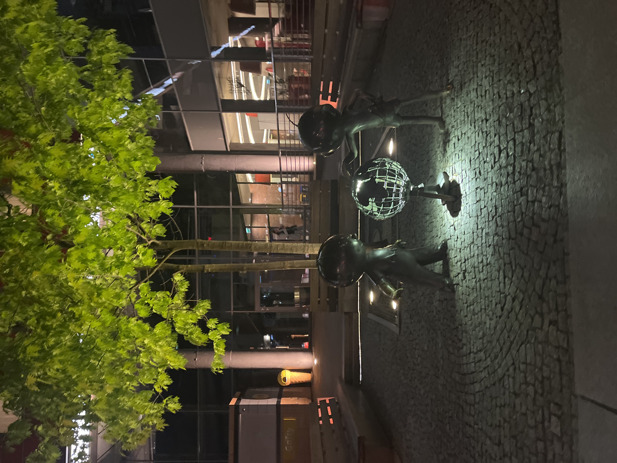 Here's a detailed description of the image:
**Overall Scene:**
The image captures a night scene in what appears to be a public square or open space in an urban setting. The orientation is vertical. The image is lit by artificial light, casting highlights and shadows.
**Subjects and Objects:**
* **Statues:** The primary subjects are two human-like statues holding up a globe made of metal mesh. The statues appear to be made of a dark material, possibly bronze.
* **Building:** A modern building with glass windows and multiple stories forms the backdrop. Light emanates from within the building.
* **Tree:** A tree with lush green foliage occupies the left side of the frame.
* **Pavement:** The ground is covered in cobblestones or paving stones.
* **Streetlight:** A street light can be seen.
**Actions and Interactions:**
The statues are posed to support the globe, creating a central focal point. The lighting creates a contrast between the sculptures and their surroundings, drawing attention to the statues.
**Environment:**
The environment is urban, with a mix of natural elements (the tree) and built structures (the building and pavement). The presence of artificial light suggests it is after dark.
**Location:**
Based on the scene, it's difficult to pinpoint an exact location. More information would be needed to discern the city and country.