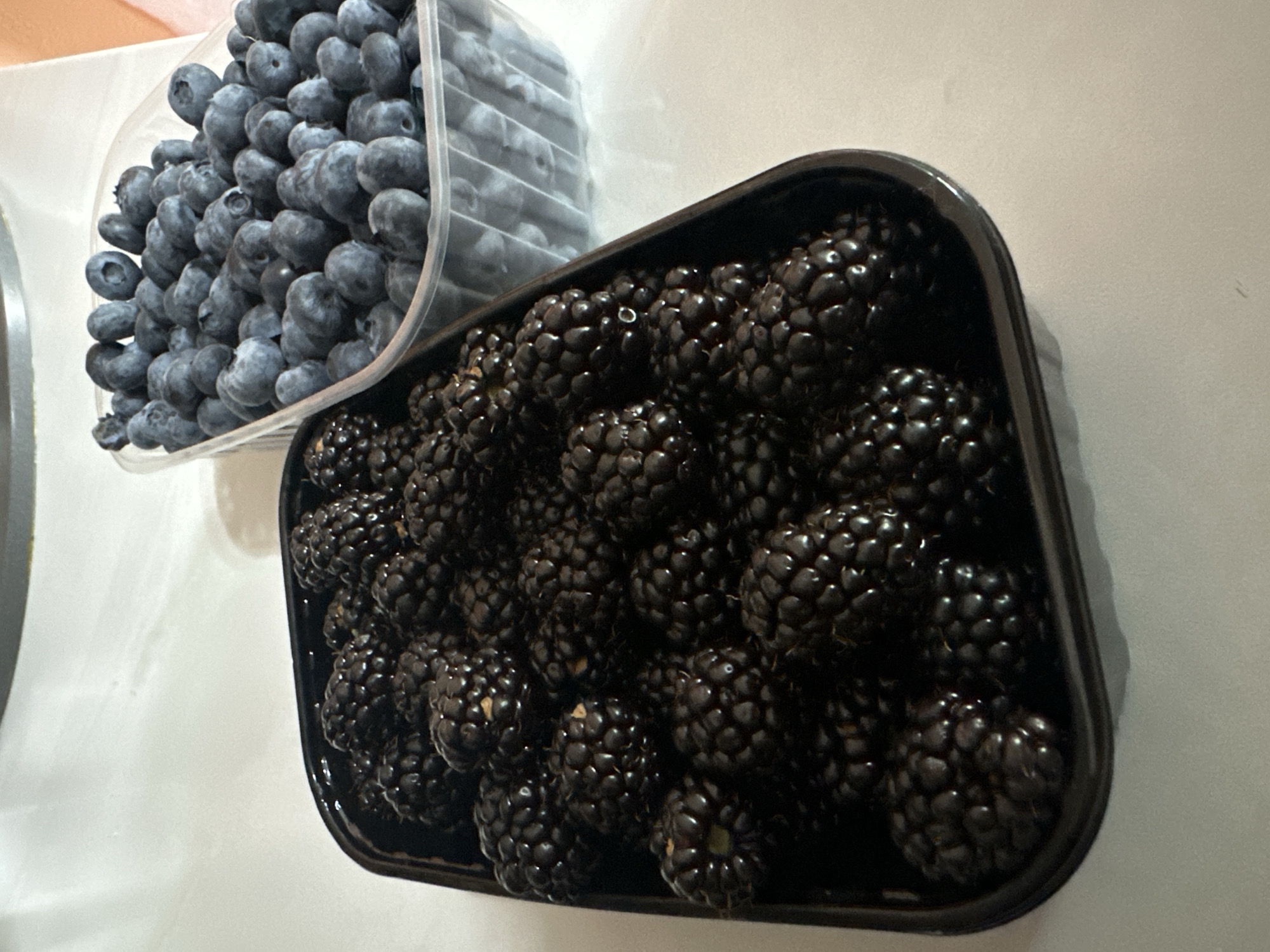 A close-up, eye-level shot shows two containers of fresh berries against a light-colored, textured surface. The container on the left is a clear plastic clamshell box, tilted and overflowing with plump, deep blue blueberries. The berries are densely packed, with some spilling over the edge. Their surface appears smooth and slightly dusty.

To the right, a black plastic rectangular container is filled with blackberries. These berries are larger and darker than the blueberries, with a bumpy, segmented surface that glistens, suggesting they are ripe and possibly slightly moist. They are also densely packed, filling the container to the brim. The lighting casts subtle shadows from the containers and berries onto the background, indicating an indoor setting, possibly a kitchen counter. There are no people visible, and no specific time of day or weather cues are apparent. The scene is focused on the fresh produce. No text is visible in the image. The overall impression is one of abundance and freshness.