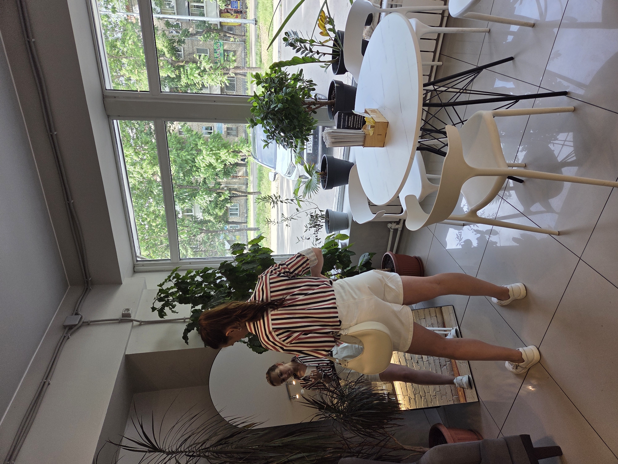 The image depicts the interior of a cafe or similar establishment in Birobidzhan, Russia. A woman with her hair in a ponytail stands facing a large, full-length mirror, wearing a striped shirt and white shorts, and carrying a cream-colored handbag. She is looking at her reflection. In the background, a large window offers a view of a tree-lined street with buildings and a silver car. Inside the establishment, several potted plants are visible, as well as a white round table with black metal legs, surrounded by white chairs. The floor is tiled. The overall impression is of a bright, casually decorated space.