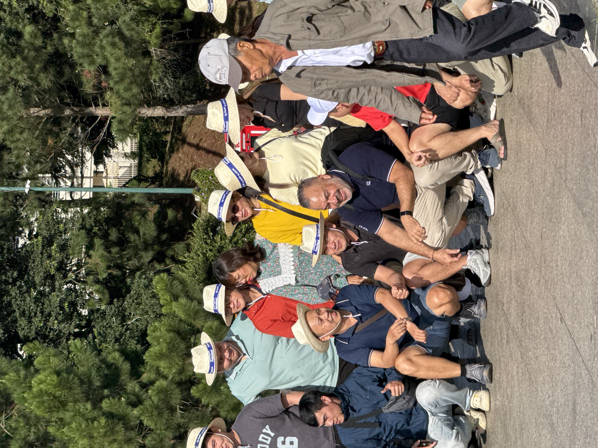 The image depicts a group of approximately ten adults, appearing to be tourists, posing for a photograph outdoors. Most of them are wearing straw hats with blue bands, and several have on sunglasses. They are gathered on a paved area, possibly a promenade or walkway, with lush green trees and foliage forming the background. In the upper right portion of the image, a white building with some fencing is visible, suggesting an urban or park-like setting within a city. The people are positioned at various levels, some standing and others crouching, all facing the camera. The atmosphere appears casual and relaxed, typical of a tourist group outing. The presence of a building and manicured greenery suggests a location like Xuan Huong Lake in Da Lat, Vietnam, known for its scenic beauty and tourist attractions.