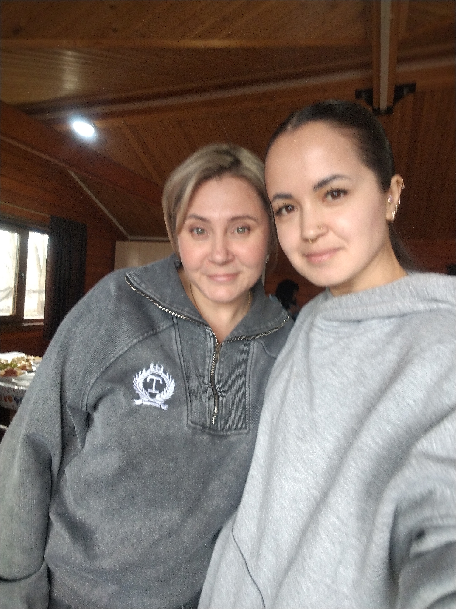 Two women are posing for a selfie indoors. The woman on the left has blonde hair, a grey zip-up sweatshirt with a white emblem on the chest, and is standing slightly behind the woman on the right. The woman on the right has dark hair pulled back, is wearing a light grey sweatshirt, and has a nose ring and earrings. The background shows a wooden ceiling and walls, a window with a dark curtain, and part of a table with food. Based on the available information, it is impossible to confirm that this image was taken in the city of Chaikovsky, Russia.