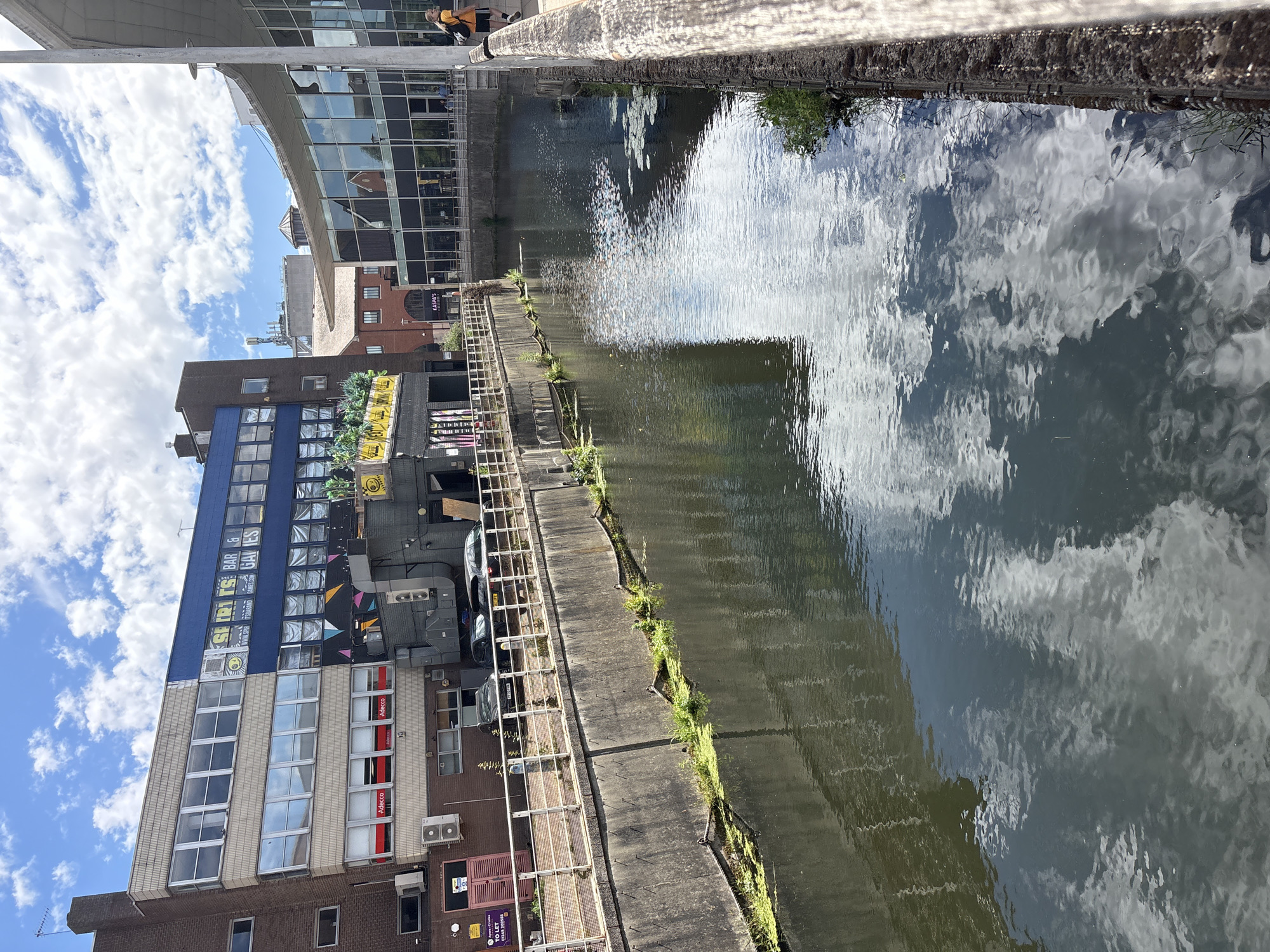 The image shows a canal with buildings on one side and a walkway along the bank. The water reflects the sky and clouds, creating a rippling pattern. The buildings are of various styles, with some featuring large glass windows and others displaying colorful murals or advertisements. One building has prominent signage that reads "SHORTS BAR & GAMES". Another building has "Adecco" advertisements. There are also some cars parked on the walkway. The sky is blue with scattered white clouds, suggesting a bright and possibly warm day. The scene appears to be in an urban environment. There are some people visible in the background, one of whom is wearing a yellow shirt. The overall impression is a busy urban waterway on a clear day.
