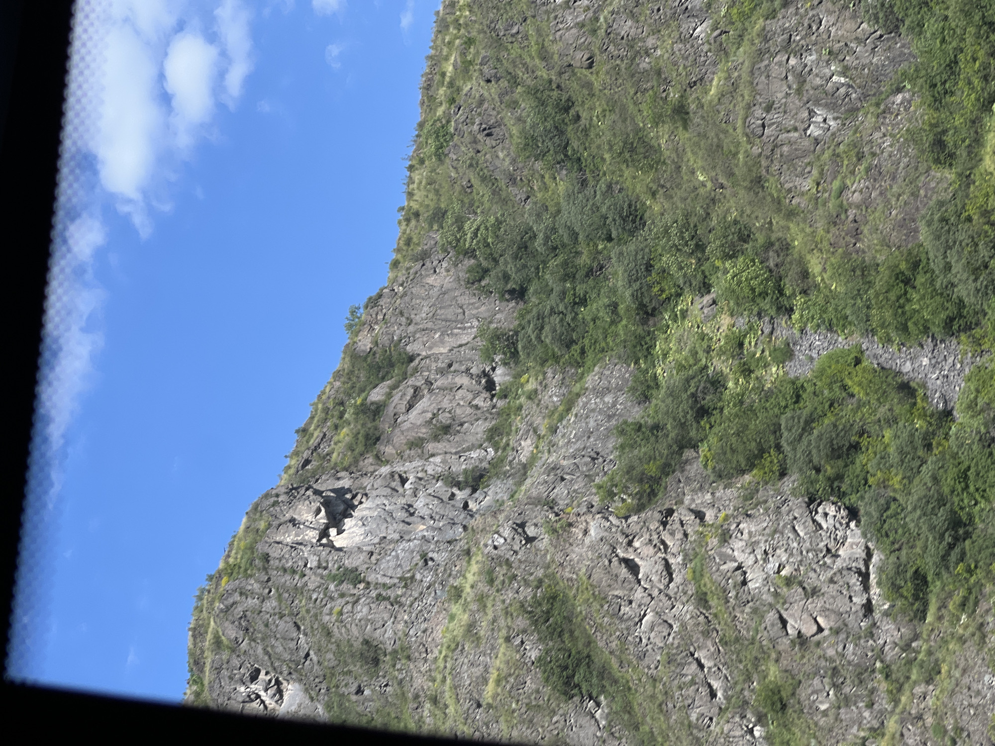 The image depicts a steep, rocky mountainside covered with green vegetation, under a clear blue sky with scattered white clouds. The foreground is partially obscured by a dark, blurry frame, possibly from a window. The rock face is rugged and textured, with visible layers and crevices. Patches of grass and shrubs grow on the slopes, interspersed with clusters of trees. The overall scene suggests a natural, undeveloped landscape. The specific location is identified as the city of Odzun, Armenia, known for its mountainous terrain and historical sites.