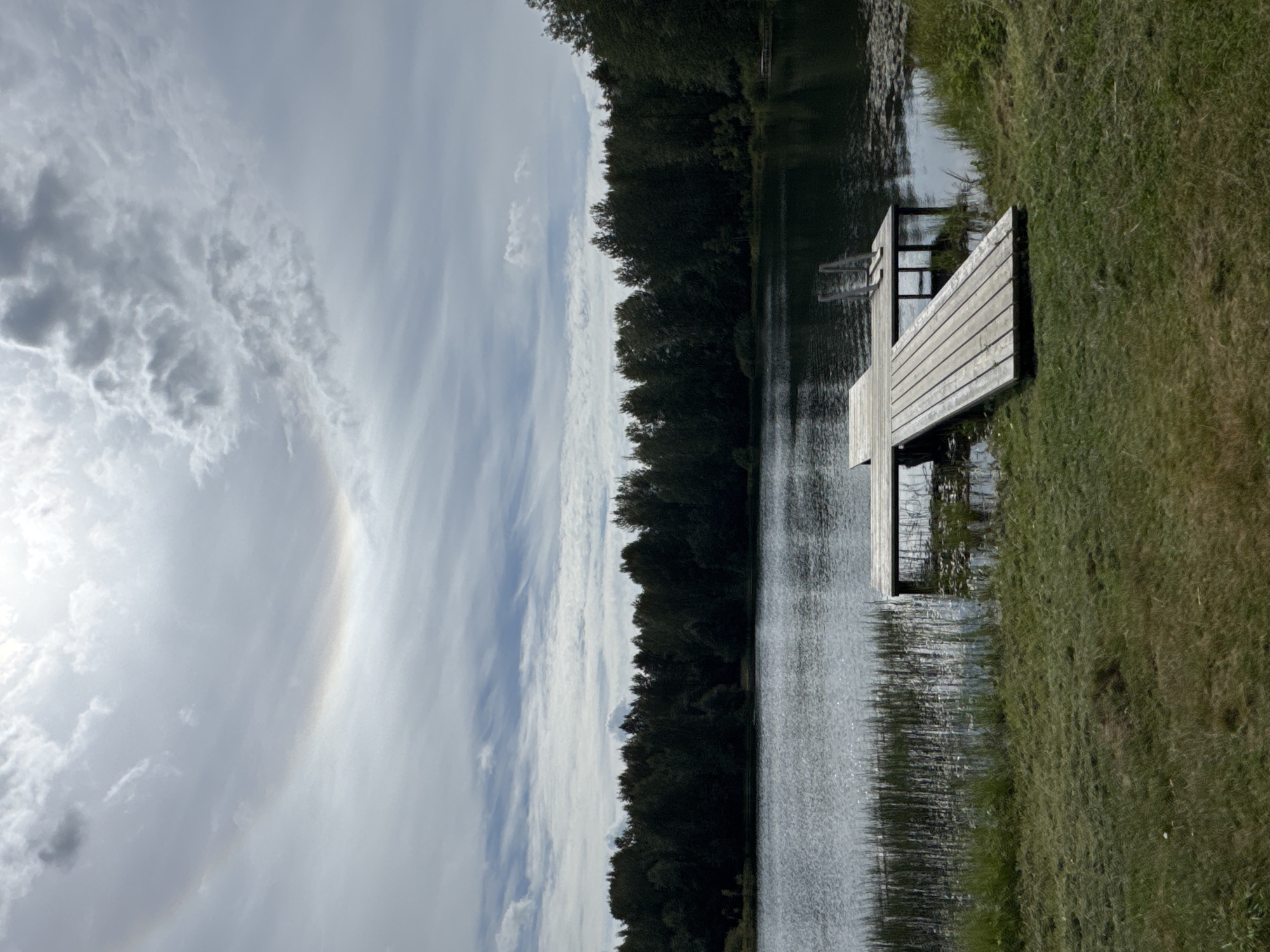 The image depicts a serene lakeside scene. A wooden dock extends into the dark, rippling water, its planks weathered and grey. The lake is bordered by a dense, dark green forest of trees. In the foreground, a grassy lawn slopes down to the water's edge, with patches of cut grass visible. The sky above is a mix of white and grey clouds, with a faint, colorful arc-like phenomenon, possibly a sun dog, visible in the upper left quadrant. The overall atmosphere is calm and natural. The image does not contain any people or actions. This scene is not located in the city of Mars, Russia.