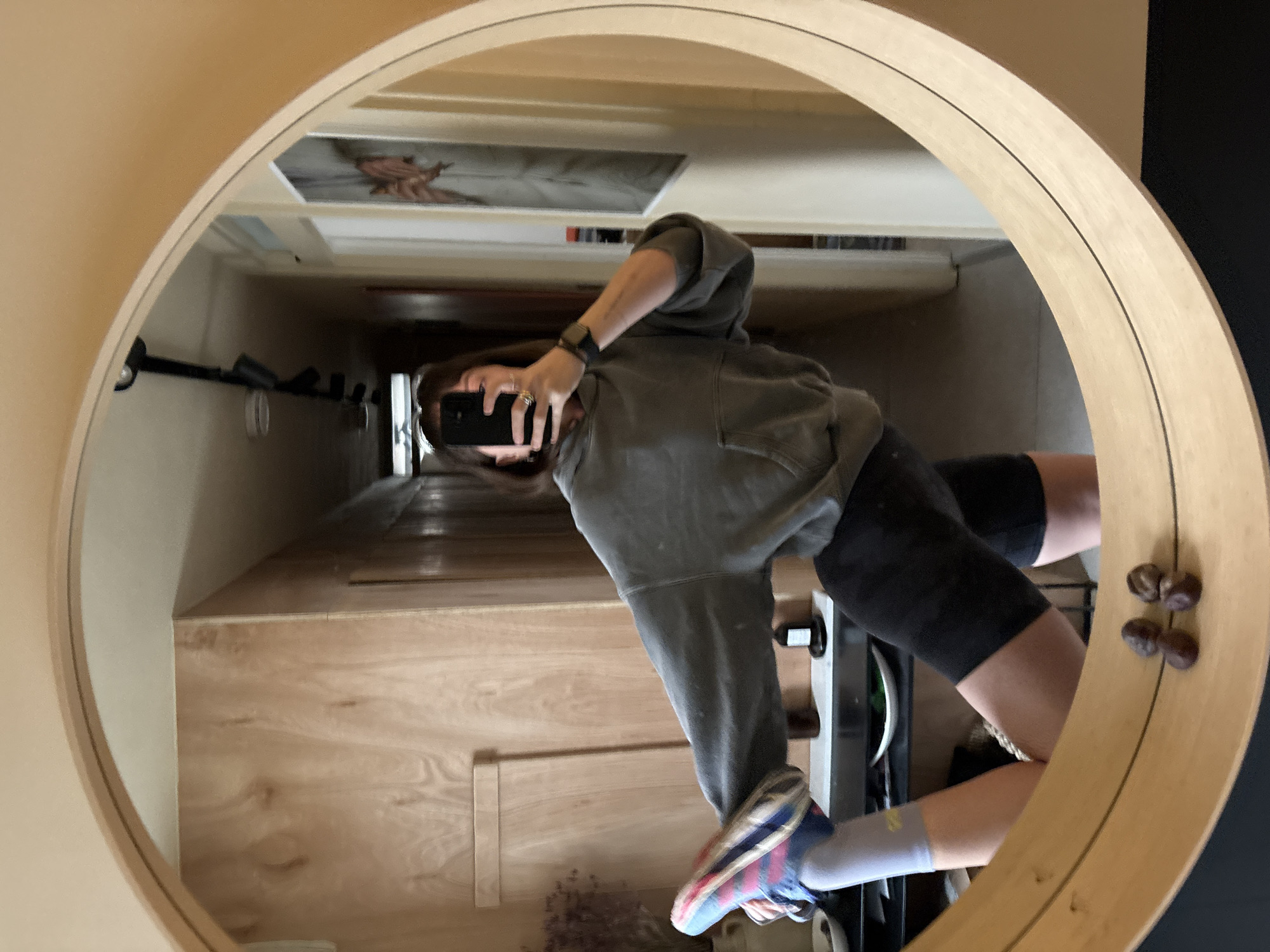 The image, rotated 90 degrees clockwise, captures a mirror selfie taken by a person standing in an indoor setting. The large, circular mirror has a wide, light-toned wooden frame, accented by a thin dark line running along its inner edge.
The subject is wearing a grey hooded sweatshirt, black athletic shorts, light grey socks, and multi-colored athletic shoes (blue, pink, and red). Their face is mostly obscured by a black smartphone, held up to take the photograph. A black smartwatch, resembling an Apple Watch, is visible on their left wrist, and a ring adorns their finger.
The mirror's reflection reveals an interior space, likely a hallway or landing with a wooden staircase. The stairs feature light wooden treads and risers, appearing to lead upwards or downwards into a darker area. A light-colored wall, possibly beige, runs alongside the staircase. On the wall above the stairs, a partially visible piece of artwork or a poster depicting a classical-style figure can be seen. Below, a small plant or decorative item sits near the base of the reflected stairs. Several dark, indistinct objects are visible on a shelf or railing along the wall. On the right side of the mirror's actual wooden frame, four small, dark, round objects, resembling chestnuts, are decoratively arranged in two pairs.
The lighting suggests an indoor daytime setting. The person's posture is casual, indicative of taking a self-portrait. No discernible text is visible. This scene is located in London, United Kingdom.