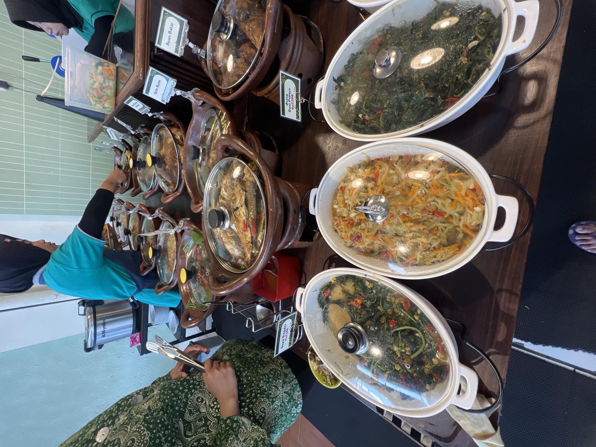 A close-up, eye-level shot captures a buffet line of Indonesian cuisine at a restaurant in Yogyakarta, Indonesia. In the foreground, three oval white dishes with clear lids are arranged on a dark wooden surface. The leftmost dish contains a dark green leafy vegetable dish with red chili peppers and corn kernels. The middle dish holds a stir-fry of shredded vegetables, including carrots and possibly cabbage, with chili peppers. The rightmost dish also contains a dark green leafy vegetable dish, similar to the one on the left, with red chili peppers. Each of these dishes is supported by black metal stands.
Behind these dishes, a row of clay pots with dark lids is visible. Some of these pots contain food items, with one pot clearly showing fried fish. To the left of these clay pots, a person wearing a green patterned dress with white paisley designs is holding a pair of silver tongs, ready to serve themselves. They are wearing a dark green long-sleeved shirt underneath the dress.
In the background, a woman wearing a teal uniform and a black hijab is serving food from a clear plastic container filled with chopped vegetables. A silver rice cooker labeled "Miyako" is visible to her left. The wall behind the buffet is painted a light blue-green color, with vertical mint green tiles above it. The floor is covered with a black mat that has a white stripe running diagonally.
Several small white signs are placed in front of the dishes, indicating the names of the food items, though they are mostly unreadable due to their size and angle. One visible sign reads "Sayur Buncis," another reads "Sate Ayam," and one reads "Ayam Bakar." The lighting suggests an indoor setting, possibly during daylight hours, with overhead lights casting reflections on the food containers. The overall impression is of a well-stocked and inviting food buffet.