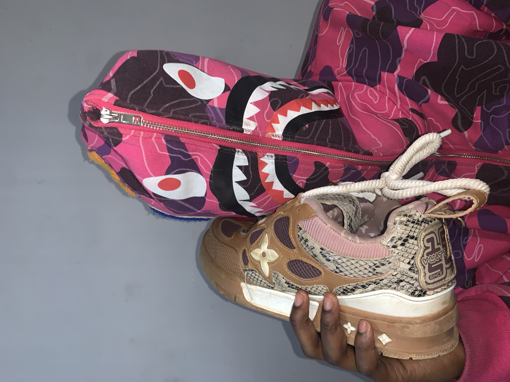A pink, purple, and brown camouflage hoodie with a shark design on the hood rests on a gray surface. The hoodie is partially unzipped, revealing a blue interior lining. A pair of Louis Vuitton sneakers with tan and brown detailing is being held by a dark-skinned hand. The sneakers feature a floral pattern, snakeskin texture, and the LV logo. The hoodie and the sneaker appear to be the focus of the image. There is no visible evidence to suggest this image was taken in Rafin Par, Nigeria.