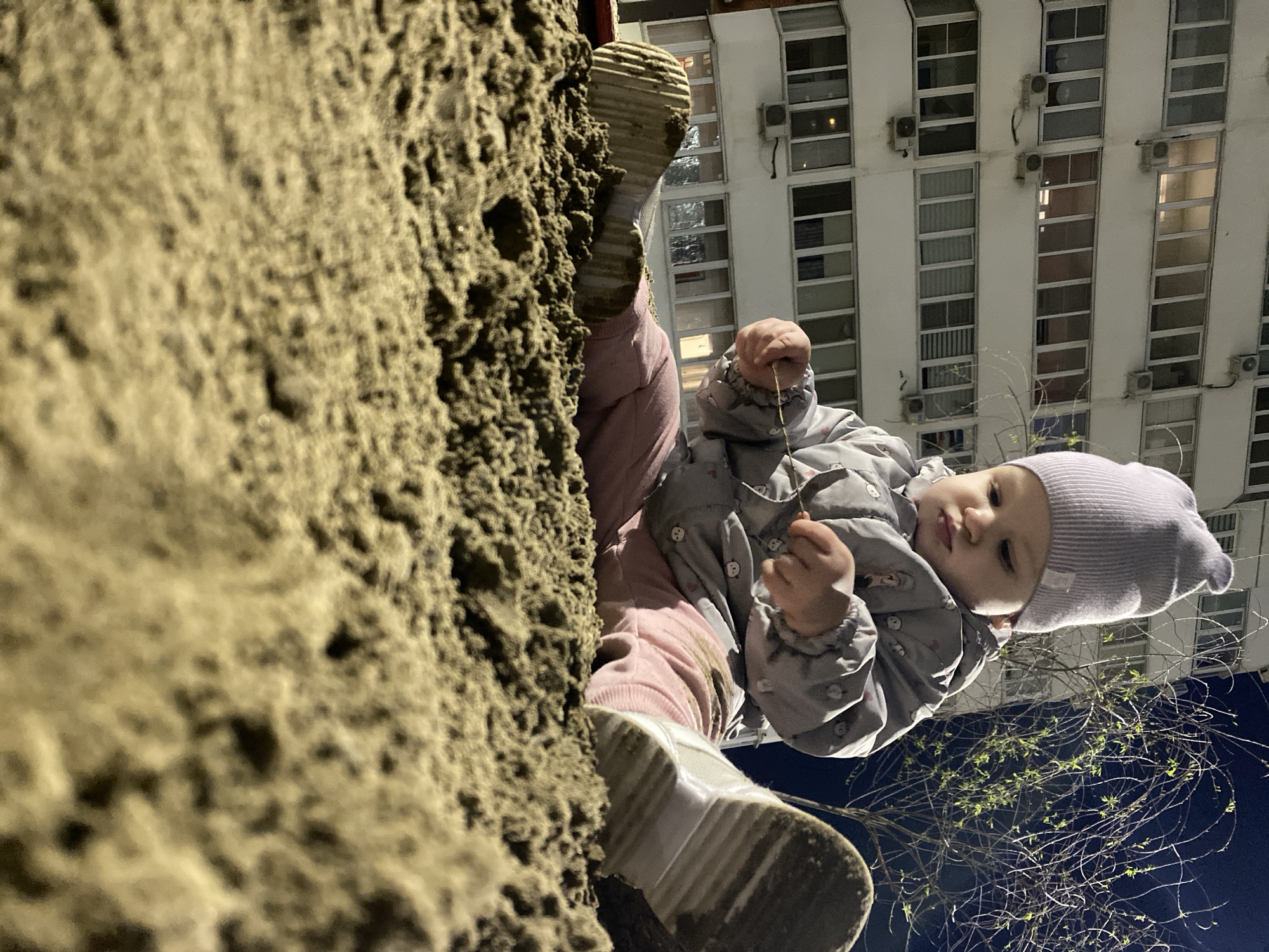 A low-angle shot depicts a child wearing a light gray beanie, a gray puffer jacket with patterns, and pink pants standing next to a large, textured tree trunk. The soles of the child's shoes are visible, appearing soiled. The background reveals a tall, white building with multiple windows and air conditioning units, suggesting an urban environment. The sky is a dark blue. The child is holding small twigs in their hands.