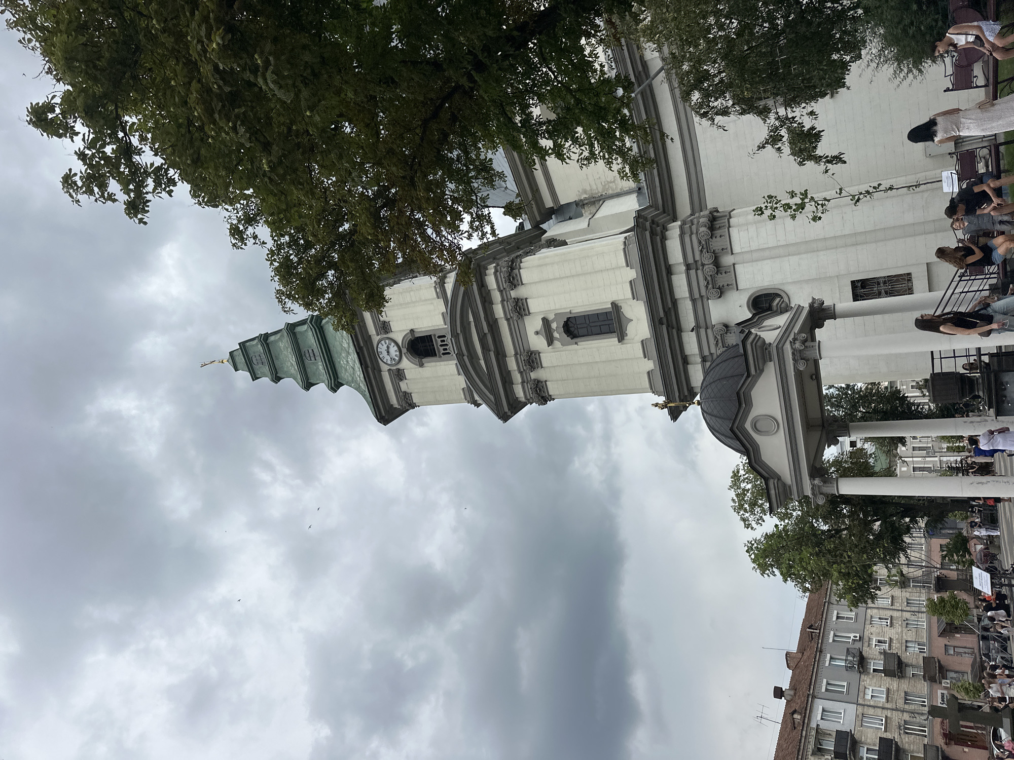 In Ternopil, Ukraine, beneath a cloudy sky, a white stone church with a green steeple and a clock tower dominates the frame. The church features ornate architectural details, including columns and decorative carvings. A dark dome-shaped structure with a cross sits at the base of the steeple. To the left, apartment buildings with multiple windows and balconies are visible. In the foreground, several people are gathered, some sitting on benches and others standing, near the church entrance. Trees with lush green foliage are scattered throughout the scene, partially obscuring the church. The overall atmosphere suggests a public space within the city.