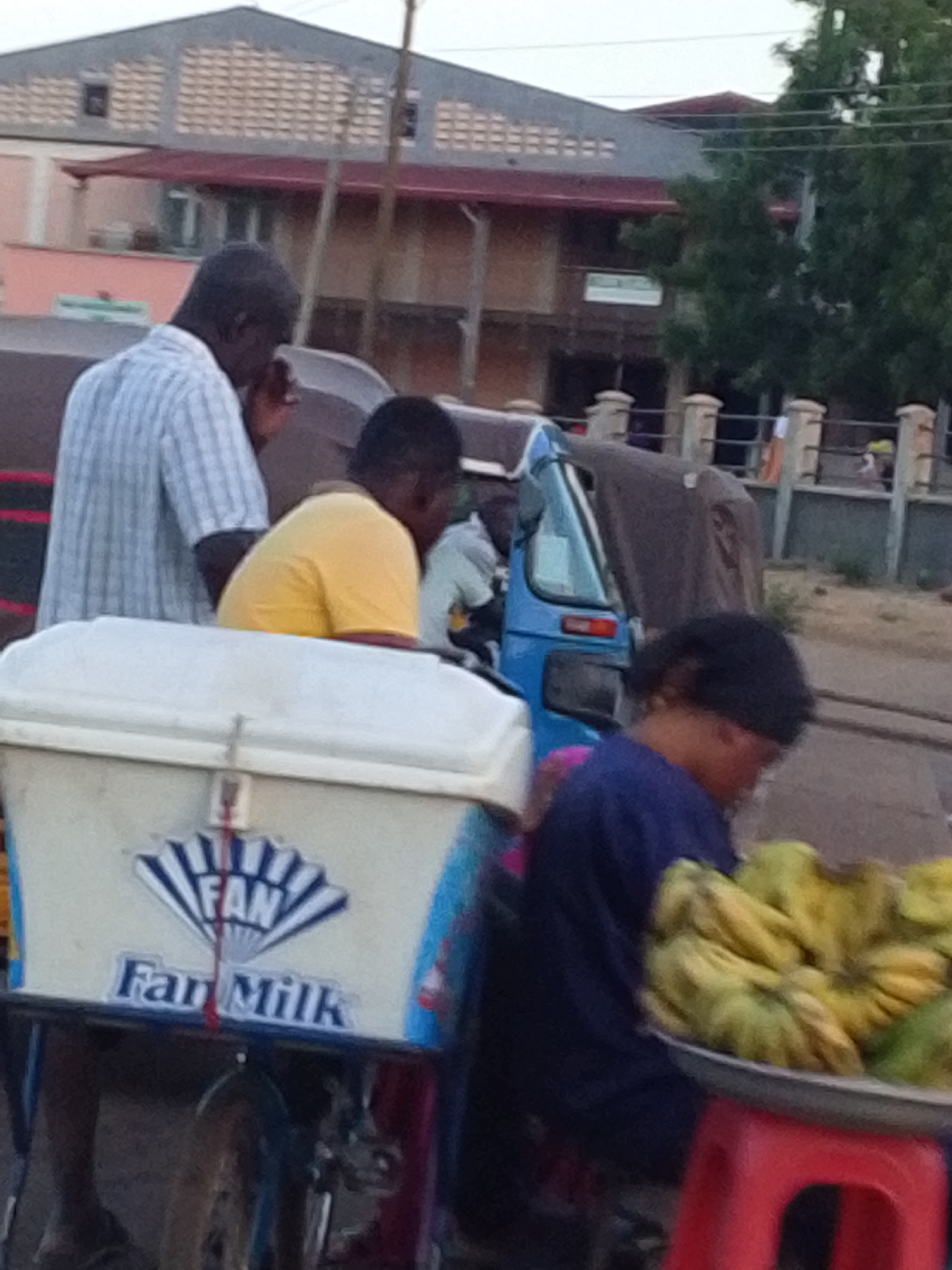 A daytime street scene in Tamale, Ghana shows several people near a blue motorized tricycle. A man in a vertically striped shirt is at the left. In front of the tricycle is a white cooler that displays "FAN Fan Milk" on its side. A woman wearing a dark shirt stands near the tricycle, in front of her, a red plastic stool is holding a silver tray with bananas. Buildings, trees, utility poles and a fence are visible in the background.