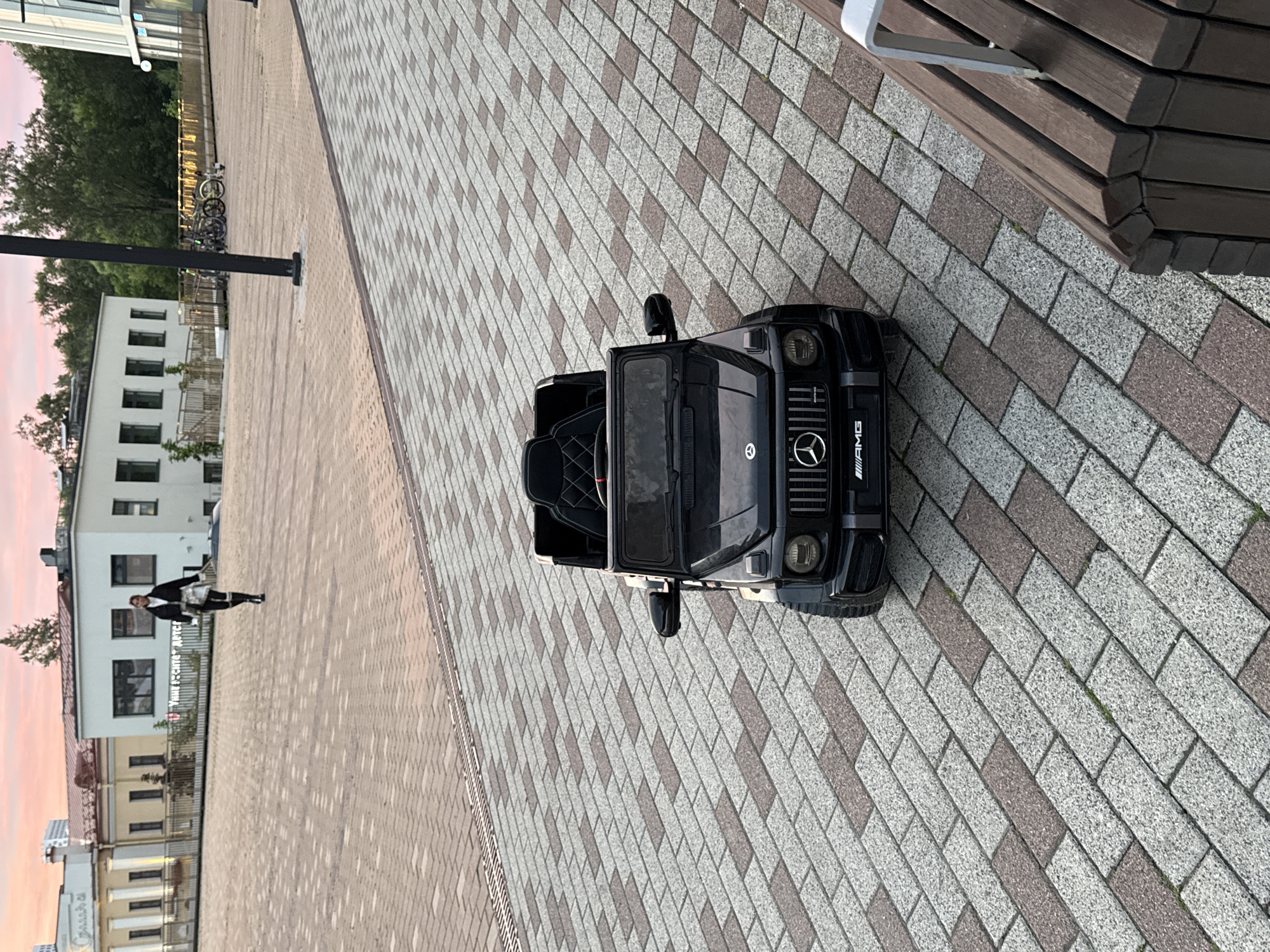 A black toy Mercedes G-wagon is parked on a patterned brick walkway in an urban plaza. In the background, a person in black clothing walks away from the camera, and behind them are modern buildings and trees. The sky is a soft pink, suggesting either sunrise or sunset. The scene appears to be in Moscow, Russia, possibly near a park or public square.
