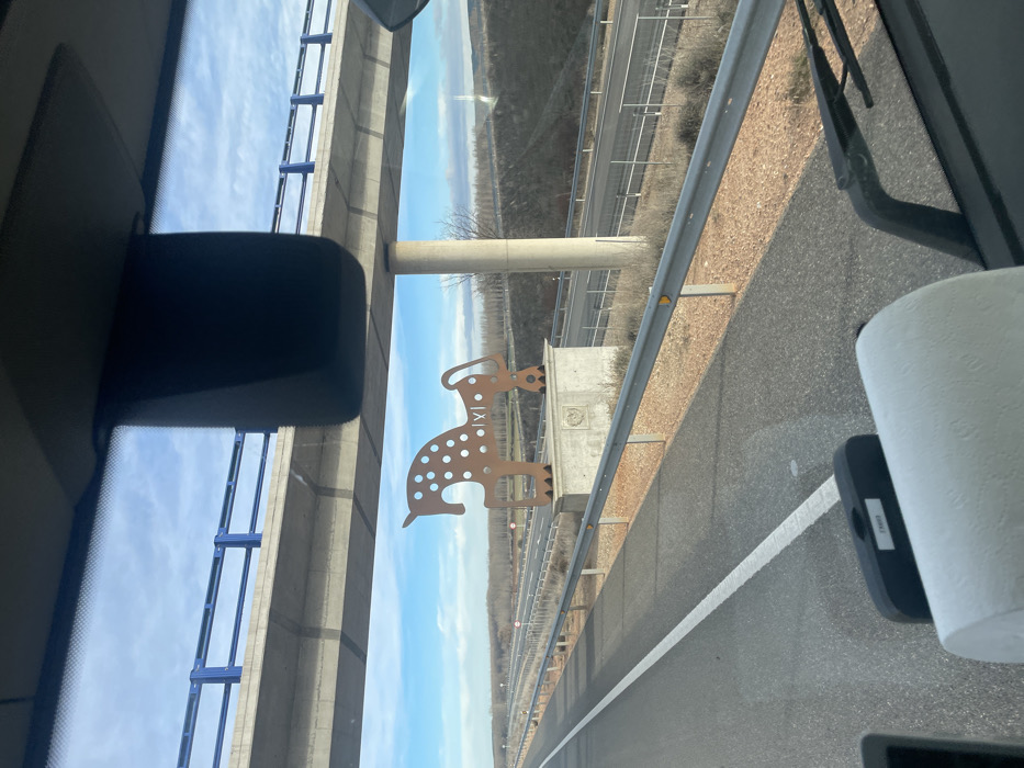 A roadside view from inside a vehicle features an overpass and a highway. A cutout dog figure with spots and Roman numerals is attached to the overpass barrier. An empty highway stretches into the distance, framed by dry landscape on either side. A roll of paper towels and a sun visor are inside the vehicle. The sky is blue with some clouds.