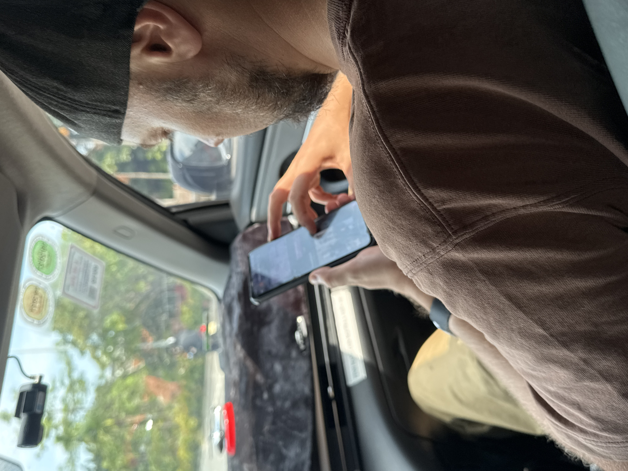 The photo shows a person in a car, likely a taxi, holding and looking at a smartphone. The person is wearing a dark cap, a brown t-shirt, and a wrist-mounted device like a smartwatch. The person has short gray hair and stubble.
Outside the car window, there is a view of a city street with trees and other vehicles. Based on the plate sticker and license plate of the other vehicles on the road, it can be concluded that the location of this photo is Hà Nội, Vietnam. A dashcam is affixed to the window.