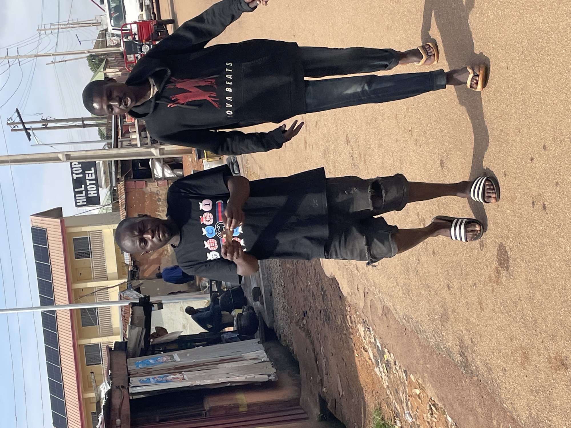 Two young men walk side-by-side on a dirt road in Jos, Nigeria. The man on the left wears a black t-shirt with colorful graphics and black shorts. He is holding some light-colored paper in his hands. The man on the right wears a black hoodie with a red graphic and the text "NOVA BEATS" in white lettering. He also wears dark blue jeans. Both men are wearing sandals. The background features a yellow building with solar panels on the roof, a sign that reads "HILL TOP HOTEL," and some corrugated metal structures. Electrical poles and wires are visible in the sky. A red tricycle and a white van are parked on the side of the road in the distance. The ground is dry and dusty, with some scattered debris. The sky is clear and blue.