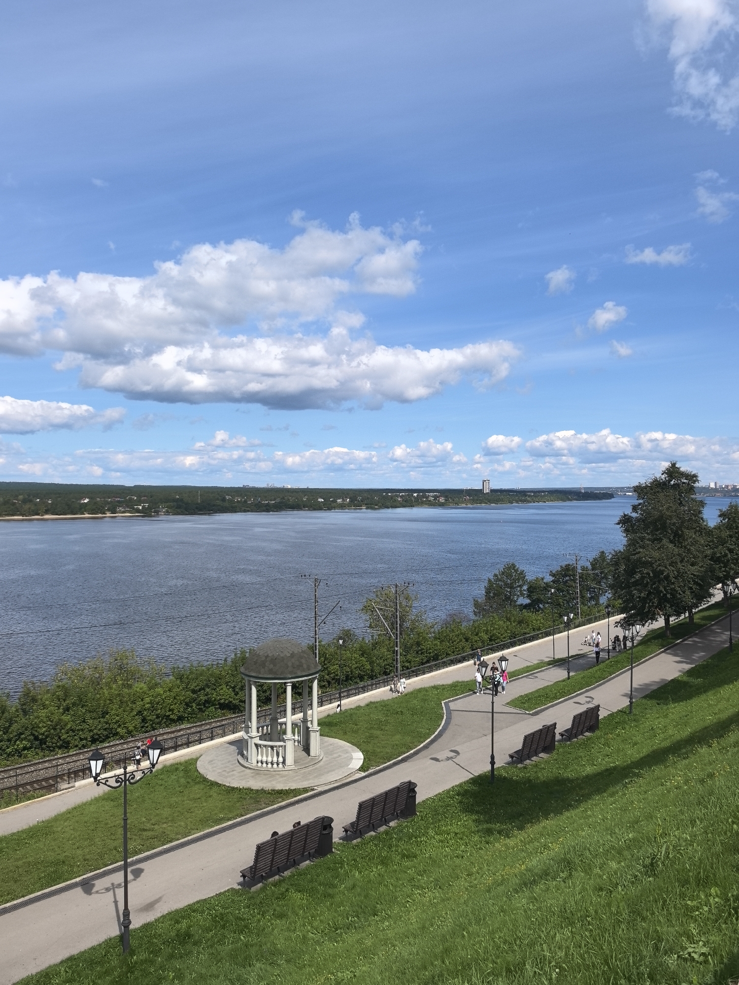 This is an eye-level, medium-shot photograph of the Perm embankment overlooking the Kama River on a sunny day. The sky is a bright blue with scattered white clouds. The Kama River fills the middle ground of the image, its dark blue water stretching to the horizon. On the far bank of the river, a tree line is visible, dotted with buildings and a tall, slender structure that appears to be a skyscraper or a communication tower.
The foreground features a well-maintained park area. A paved pathway curves through the scene, lined with green grass on either side. Several dark wooden benches are placed along the path, some occupied by people. A tall lamppost with two lights stands prominently on the left side of the pathway, casting a shadow on the grass. Further along the path, a white gazebo with a dark roof stands on a circular paved area. People are seen walking along the pathway, some in groups, enjoying the pleasant weather. Lush green trees and bushes line the edge of the embankment, separating the park from the river. In the distance, on the other side of the river, a cityscape is visible, suggesting a populated area. The overall impression is one of a serene and attractive urban recreational space.