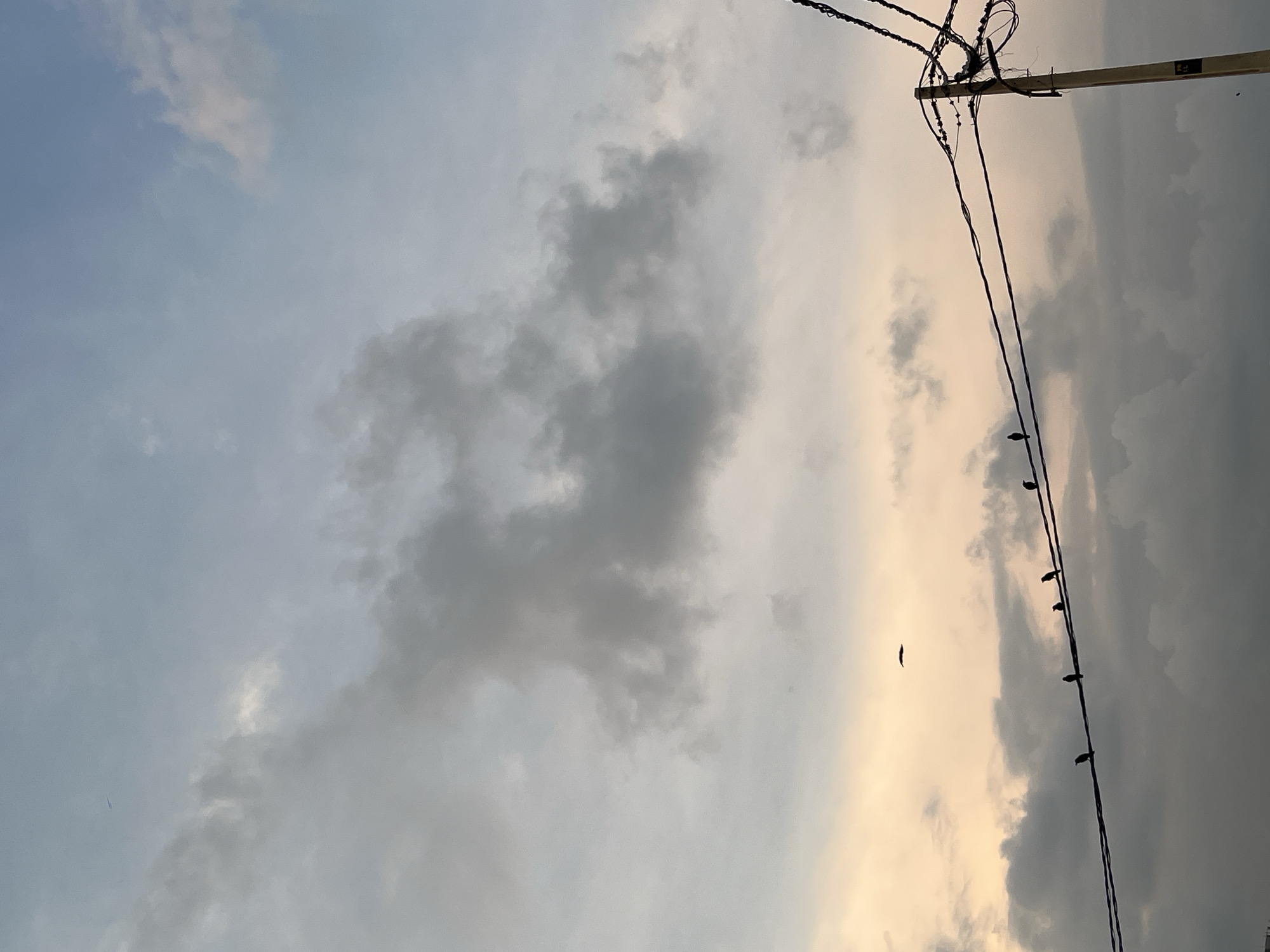 The image shows a view of the sky in Galle, Sri Lanka, with a utility pole and wires visible in the upper right corner. Several birds are perched on one of the wires. The sky is filled with clouds, ranging from light blue to dark gray.
