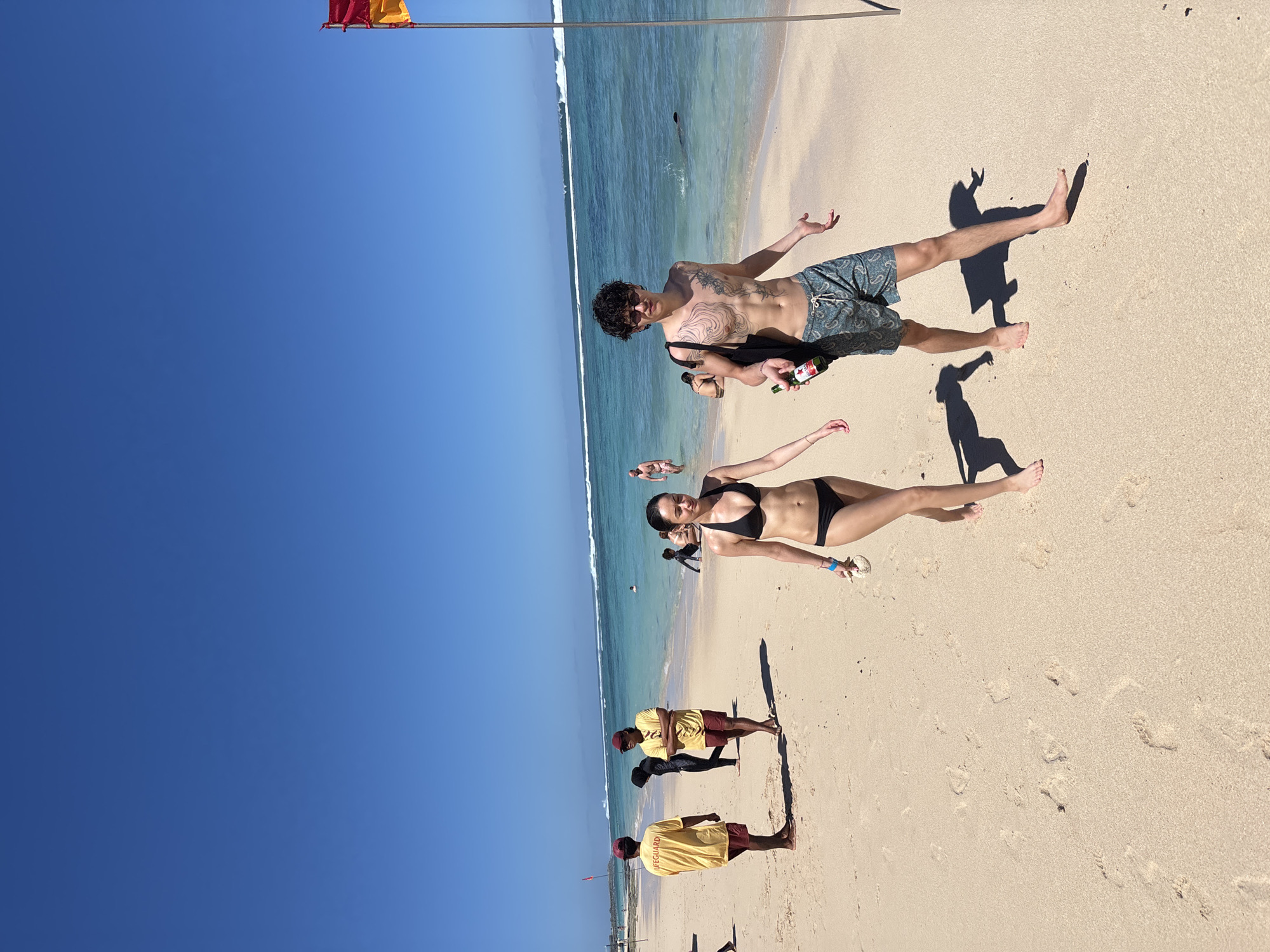 The image captures a sunny day at a beach in Kuta Selatan, Indonesia. The sky is a clear, vibrant blue. The ocean is turquoise with white waves breaking near the shore. The sand is light beige and shows footprints.
A man and a woman are standing on the beach. The man has dark curly hair, a tattoo, and is wearing patterned board shorts. He is holding a black bag and two bottles of alcohol. The woman is wearing a black bikini.
Further down the beach, there are three lifeguards wearing yellow shirts with "LIFEGUARD" printed on the back and maroon shorts. They are standing close together.
In the distance, there are other people in the water and on the sand. A flag is visible, red and yellow, attached to a metal pole, indicating the swimming conditions.