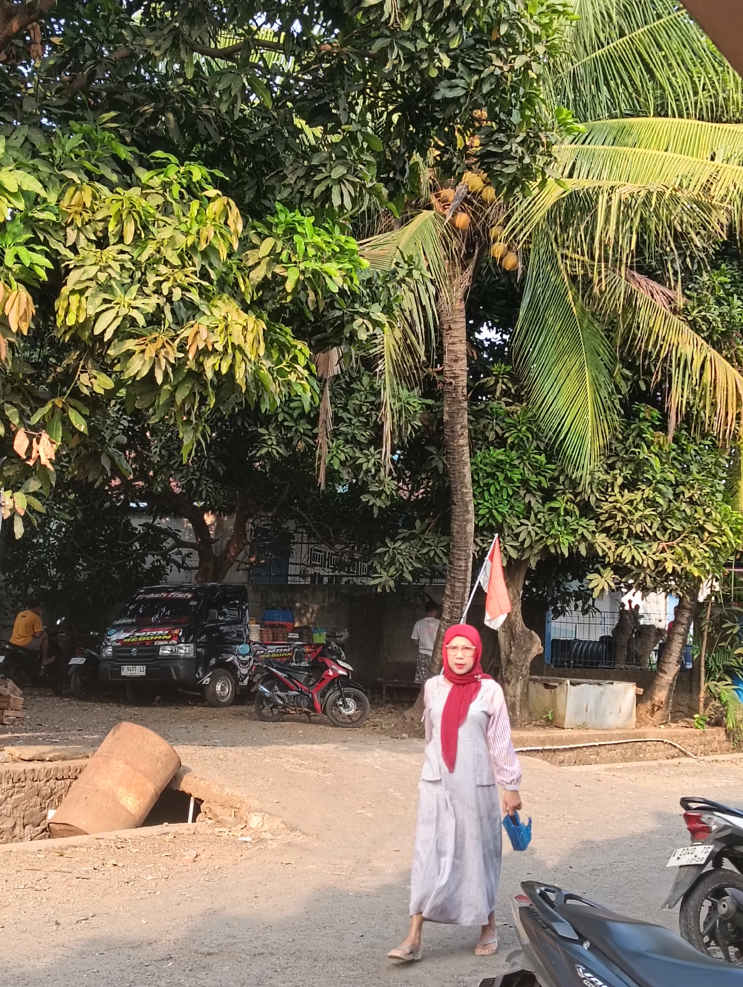 A woman wearing a red hijab and a long, gray and white striped dress walks on a dirt road in Cluwak, Indonesia. She carries a blue bag in her right hand. Behind her, a small Indonesian flag flies from a tree. In the background, several parked motorcycles and a black pickup truck are visible under the shade of large trees. A large, rusted metal barrel lies on its side in a pit to the left of the road. The sun is shining, casting shadows and illuminating the scene.