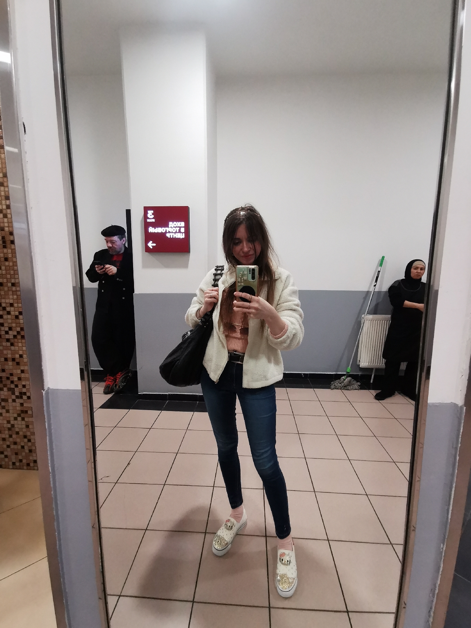 The image depicts a woman taking a selfie in a mirror. The reflection reveals an indoor public space, possibly a transit hub like a metro station or shopping center in Moscow, Russia, as suggested by the Cyrillic text on the sign in the background.
The woman in the foreground is wearing a pink top, blue jeans, and a white jacket. She has dark hair and is holding a smartphone, taking a picture of herself.
In the mirror's reflection, there are two other people: one man is wearing a black coat and beret, looking at his phone. He is standing near a red sign with white Cyrillic text and a white arrow pointing left. To the right of the frame in the reflection, there's a woman in a black hijab, standing near a white radiator with a mop resting against it. The floor is tiled, and the walls are white with a grey lower section.