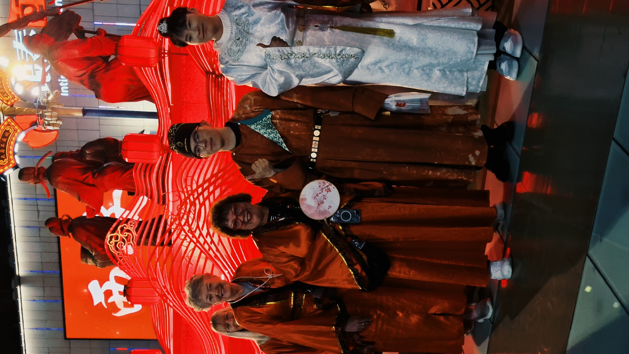 The photograph, taken in Yanta District, China, shows a group of people in traditional Chinese clothing posing in front of a red decorative display, likely for a festival or celebration. A young person in a white robe with silver trim stands at the top, next to an individual in a brown robe with a black belt. Below them, a person with curly hair is holding a fan. Further down, a woman with short, light hair stands next to another person. The background includes large red decorations and a screen. The floor is tiled.