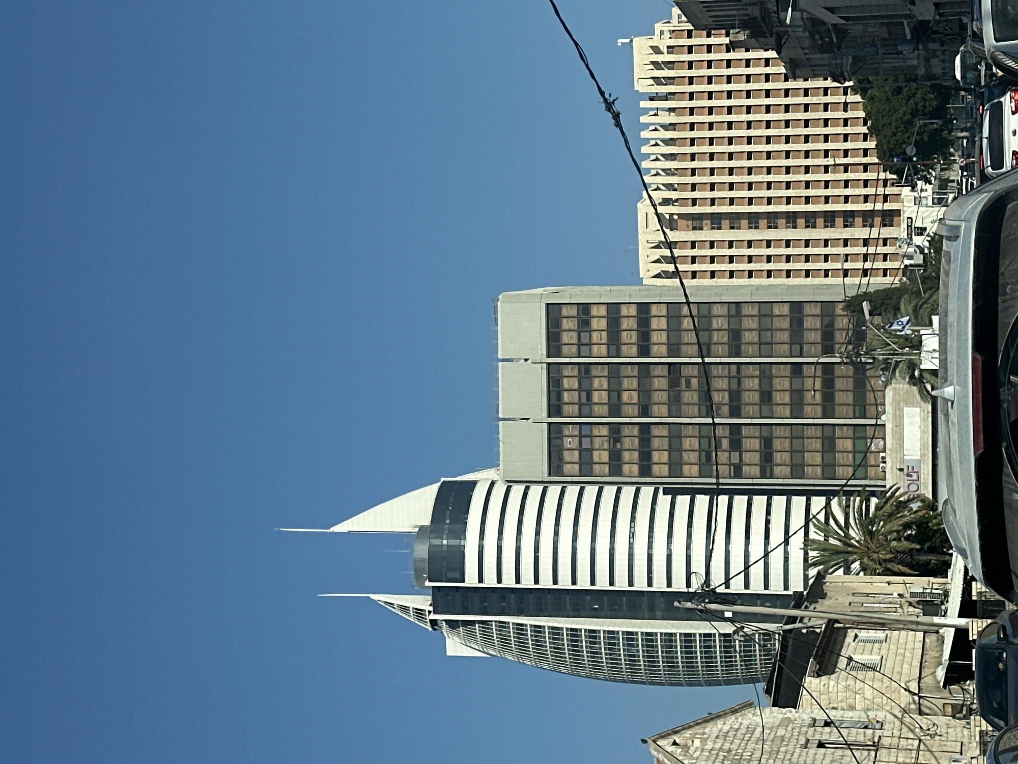 The image shows a street view in Haifa, Israel, with several modern buildings dominating the skyline. A prominent, curved skyscraper with a distinctive white, sail-like top stands to the left, featuring a facade of vertical windows. Adjacent to it is a tall, rectangular building with horizontal white bands and dark windows. Further to the right, another building displays a pattern of vertical fins and numerous small, dark windows. In the foreground, the tops of cars are visible, indicating traffic on the street. Several power lines crisscross the upper portion of the frame. The sky is clear and blue.