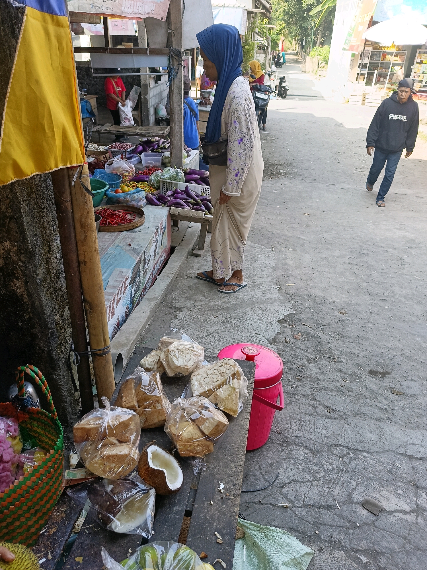This image depicts an outdoor market scene in Manisrenggo, Indonesia. In the foreground, several food items are displayed on a wooden surface. These include bags of what appears to be fried tofu or tempeh, a halved coconut, and a woven basket filled with colorful snacks. To the left, a tall yellow flag is visible, partially obscuring a market stall. Behind the stall, a person in a red shirt is visible.
In the mid-ground, a woman wearing a blue hijab and a patterned dress stands looking to the left, her hand resting on the stall's counter. Further down the paved road, another woman in a yellow headscarf is seen near a motorcycle, and behind her, a man in a dark hoodie and jeans walks away from the camera. The environment suggests a typical street market with buildings and trees lining the road in the background. A bright pink cooler is placed on the ground near the market stall.