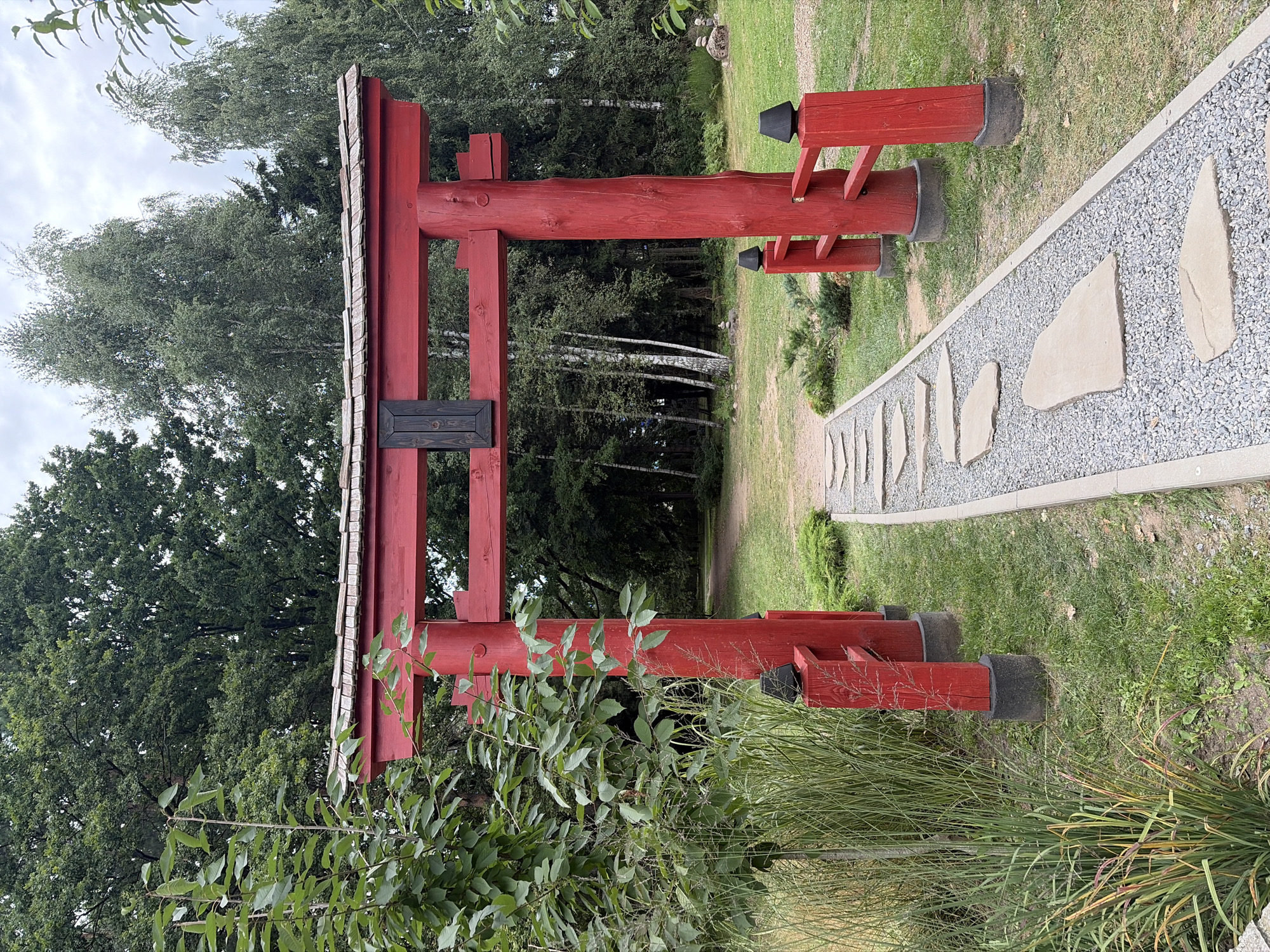 A red Japanese torii gate stands in a park-like setting, framing a path of flat stones set into gravel. The gate is made of thick wooden posts and beams, with a shingled roof. To the left of the gate, a lush green tree with a thin trunk grows, its leaves brushing against the gate. Behind the torii gate, a dense forest of trees, including birch trees with white trunks, creates a natural backdrop. The ground is covered in green grass and there are hints of other foliage scattered around. The sky above is overcast with a soft, diffused light.