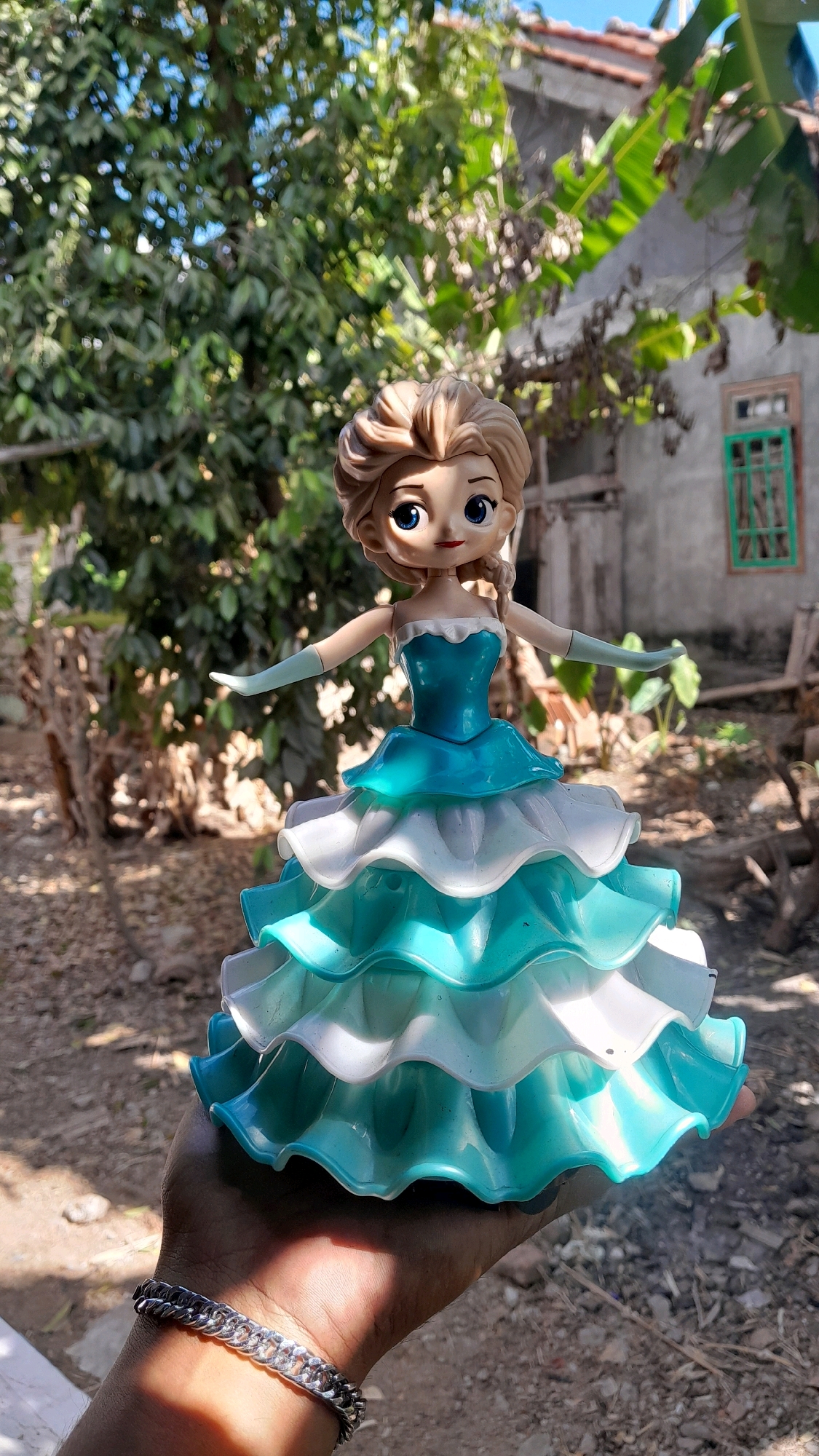 A doll, resembling the character Elsa from Disney's "Frozen", is held in a hand with a silver bracelet on the wrist. The doll is wearing a layered blue and white dress with ruffled tiers. The background shows lush green trees, a weathered building with a green-paned window, and a rustic outdoor setting. The scene appears to be an outdoor area, possibly a yard or garden, in a rural or semi-rural location. The sunlight casts shadows on the hand and the doll, indicating a bright day. The overall impression is that of a posed photograph of a toy. The presence of banana trees in the background, along with the style of the building, are indicative of a tropical or subtropical climate, consistent with locations like Binangun, Indonesia.
