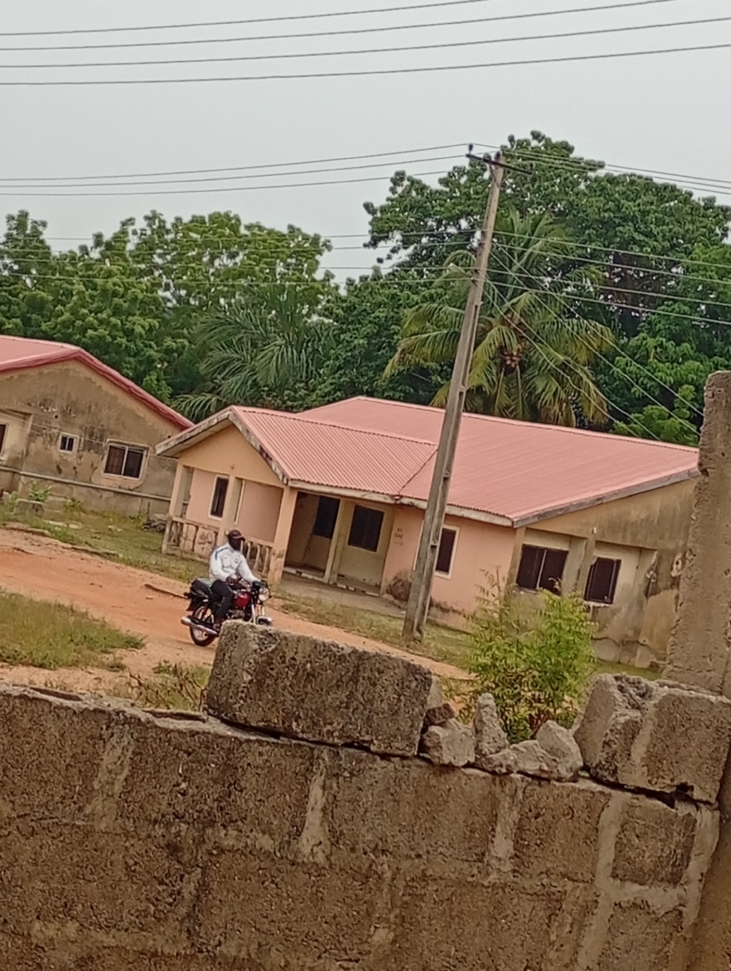The image depicts a man on a motorcycle in front of a building. The building is single-story with a red roof and light-colored walls. There is another similar building to the left, partially visible. Green trees are visible in the background and an electrical pole with wires. In the foreground is a partially constructed wall made of cinder blocks. The location appears to be Unknown, Unknown. The man is wearing light-colored clothes. He is riding the motorcycle on a dirt path. The overall environment suggests a rural or suburban setting.