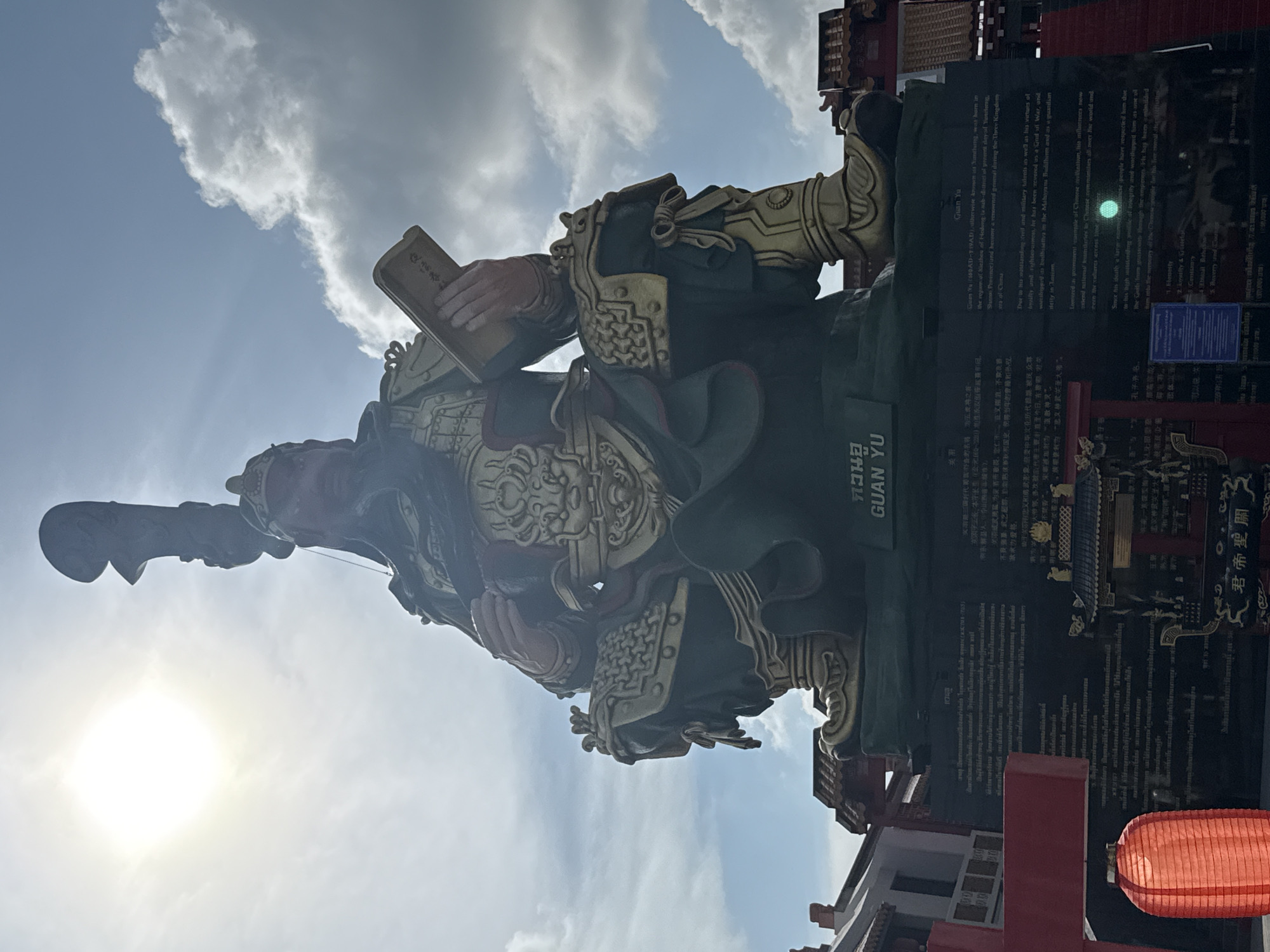 The image displays a large, ornate statue of Guan Yu, a revered Chinese general, set against a bright blue sky with scattered white clouds. The statue is richly detailed, featuring dark green robes with elaborate gold armor and red accents. Guan Yu is depicted holding a book in one hand, while a part of a halberd or spear is visible near his left shoulder, extending upwards. The sun is shining brightly in the lower left, casting a strong glare and backlighting the statue.
To the right of the statue, dark plaques with detailed inscriptions are visible. These plaques contain text in both Thai ("กวนอู") and English, identifying the figure as "GUAN YU". The English text describes him as a "renowned general during the Three Kingdom era of China" (160AD-219AD), revered as a "God of War," a "bodhisattva in Mahayana Buddhism," and a "guardian deity in Taoism" due to his "outstanding civil and military talents as well as his virtues of loyalty and righteousness." The text also lists virtues associated with him, including "Honesty," "Loyalty & Righteousness," "Generosity," and "Bravery and Courage."
In the lower right, traditional red lanterns and architectural elements with intricate dark roofs suggest a temple or cultural compound. The overall scene indicates a sunny day. This statue is located in Baan Hua Thanon, Thailand, a place known for its cultural diversity.