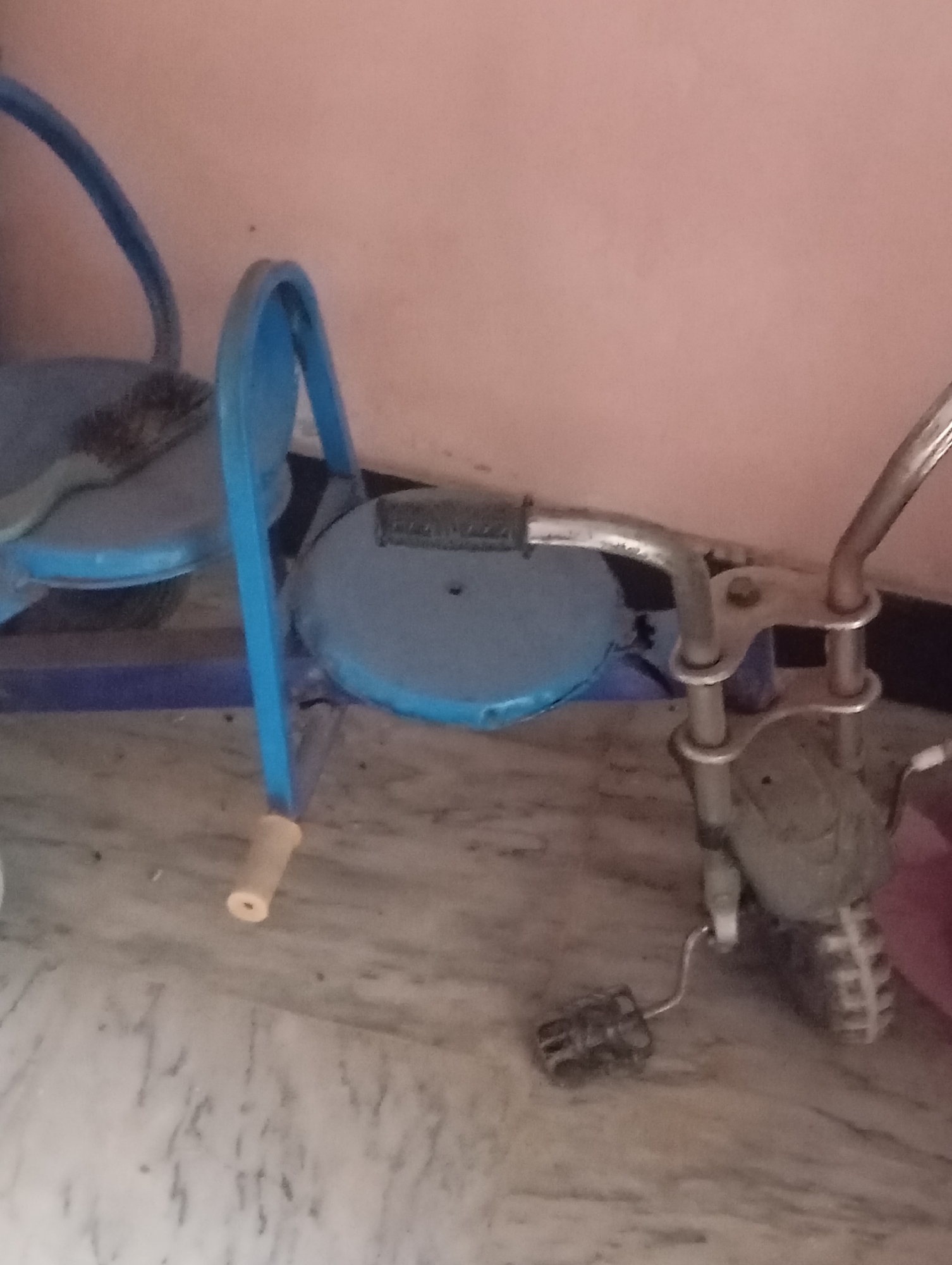 The image shows a blue tricycle with a silver handlebar and pedals. The tricycle is positioned on a floor that appears to be made of marble or a similar stone material. The background is a plain pink wall.
The tricycle itself has two blue seats, one behind the other, and a blue frame. The front handlebar is silver and has black grips. One silver pedal is visible, attached to a crank. The front wheel is partially obscured but appears to be silver or grey. There is a black grip on the left handlebar. The back of the tricycle has a visible seat and a blue frame that extends upwards.
There is no activity or event taking place; the tricycle is stationary. The lighting suggests it is daytime, but no specific time can be determined. The location is indoors, as indicated by the wall and floor. There are no people visible in the image.
Notable details include the color of the tricycle (blue and silver), the material of the floor (marble-like), and the pink wall in the background. The tricycle appears to be designed for children, possibly a tandem tricycle given the two seats. Some dirt or debris is visible on the floor and on the tricycle itself. There is no visible text in the image.