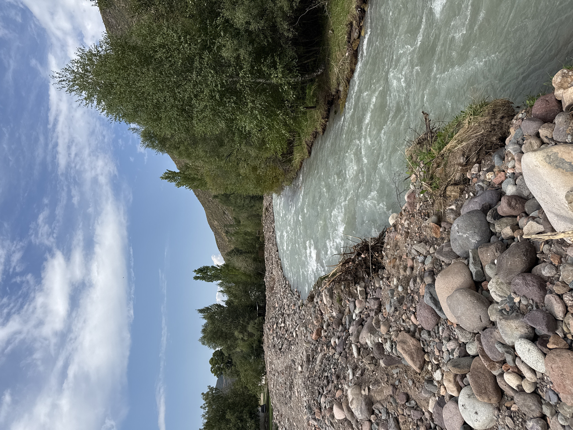 The image shows a wide, turbulent river flowing through a rocky, treed area. The river is a light, milky green color, with whitecaps indicating a strong current. The riverbank on the left is covered with a dense layer of various sized, multicolored rocks. On the right bank, a line of mature green trees grows along the slope of a barren hill. The sky above is blue with scattered white clouds. In the distance, more trees and hills are visible. The scene appears to be a natural landscape in a mountainous or hilly region, consistent with the area around Leskhoz, Kazakhstan. There are no people or animals visible in the image.