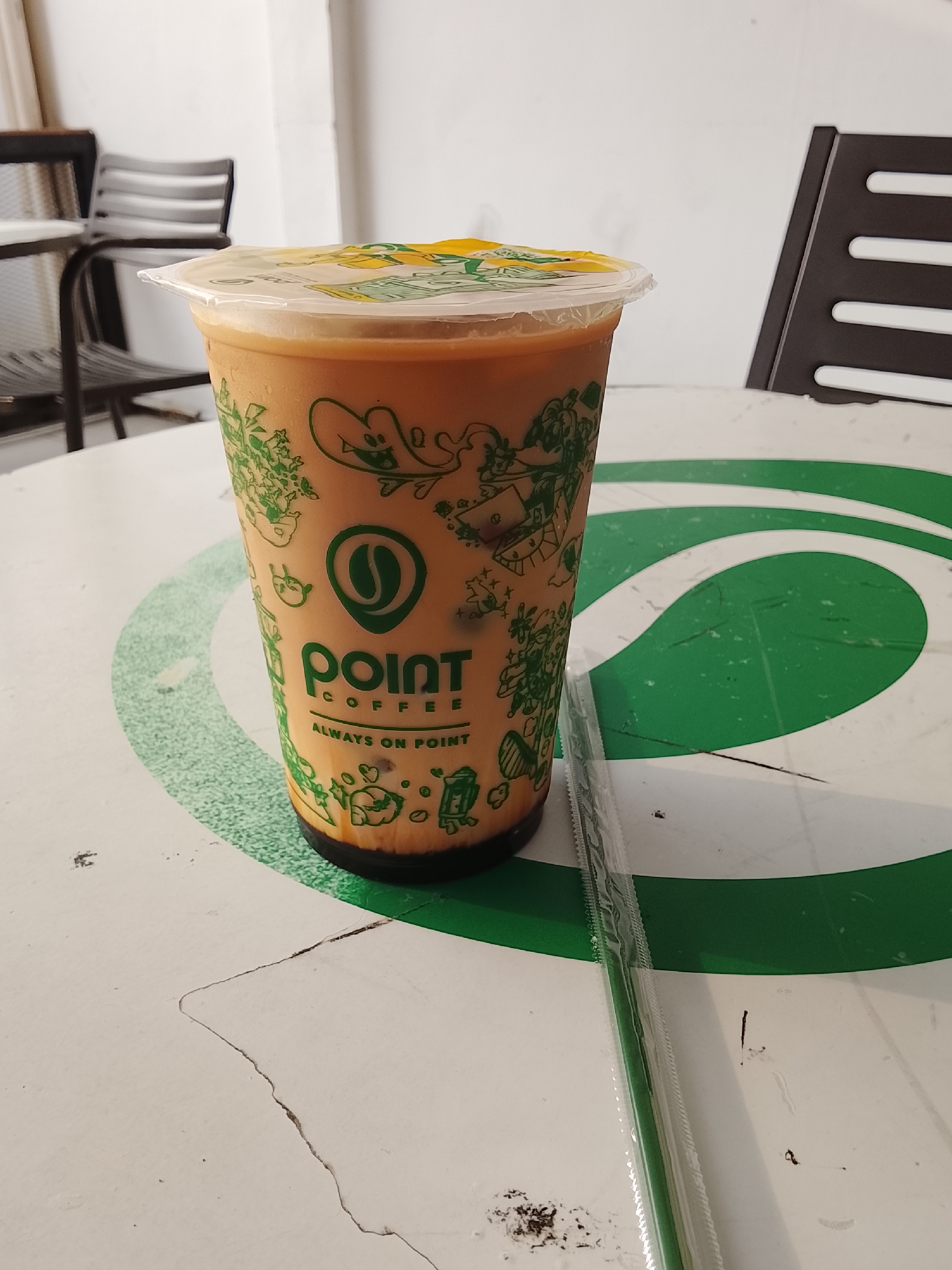 A clear plastic cup filled with a light brown, iced beverage is situated on a white table with a green circular design. The cup has a clear plastic lid with a yellow insert and features green graphics, including the "point COFFEE ALWAYS ON POINT" logo with a coffee bean icon. A green, individually wrapped straw lies to the right of the cup. In the blurred background, two dark chairs with metal frames and slats are visible against a white wall. The setting appears to be an indoor cafe or dining area, possibly in Kedungwuluh, Indonesia, given the context. The table surface shows some wear and tear, including scratches and a noticeable crack.