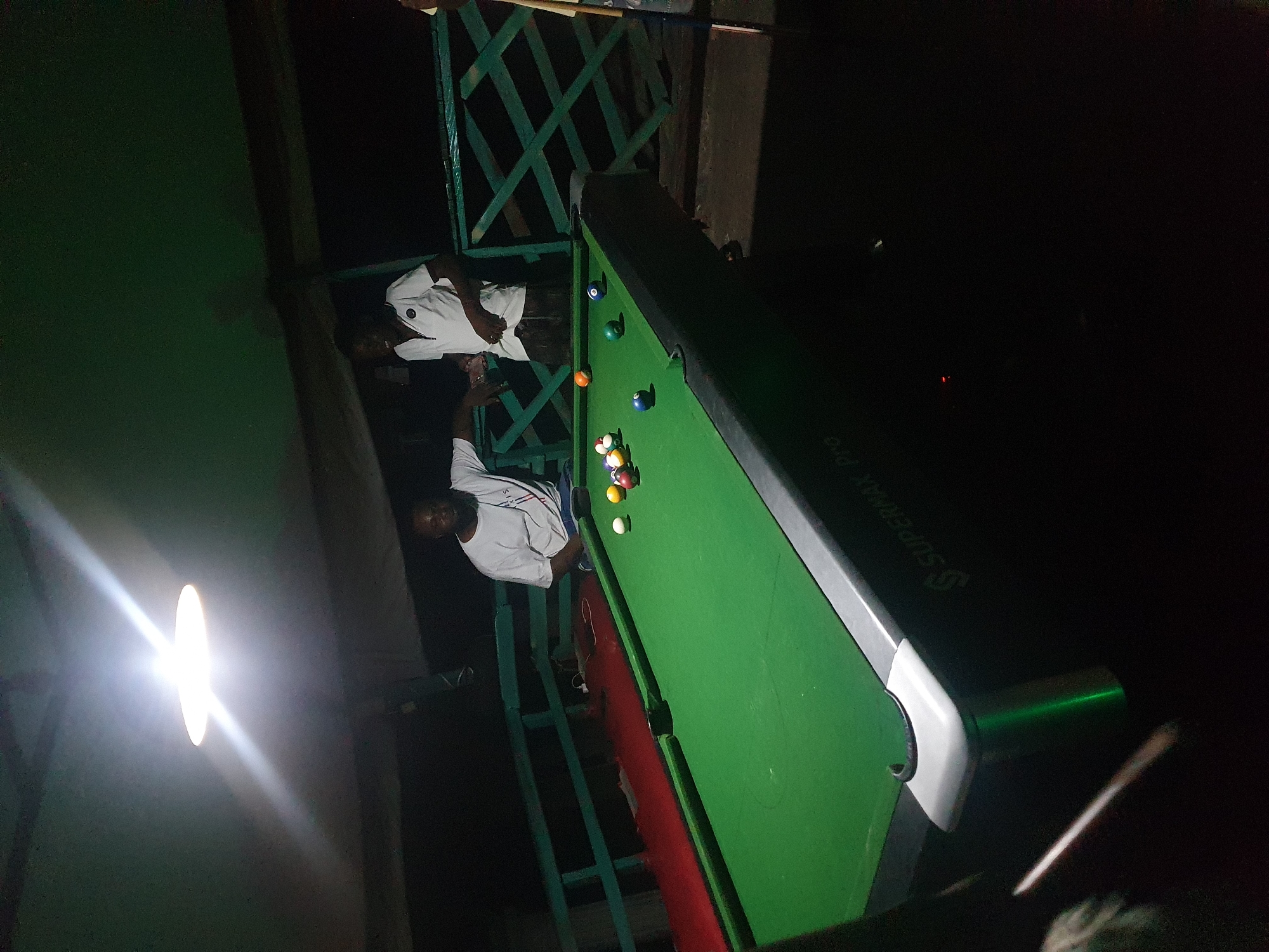 The image depicts an indoor pool hall, likely located in Lokoja, Nigeria, based on the context provided. Two men are present, standing near a green pool table. One man, wearing a white t-shirt, is leaning against a railing, while the other, also in a white t-shirt, is standing and looking towards the table. Several pool balls are scattered on the table, suggesting a game is in progress or has recently concluded. The lighting in the room is dim, with a bright overhead lamp casting a strong glare on the ceiling. The environment appears to be somewhat rustic, with visible wooden structures and a railing that suggests an elevated or open-air setting. The pool table itself has a "SUPERMAX Pro" logo visible on its side.