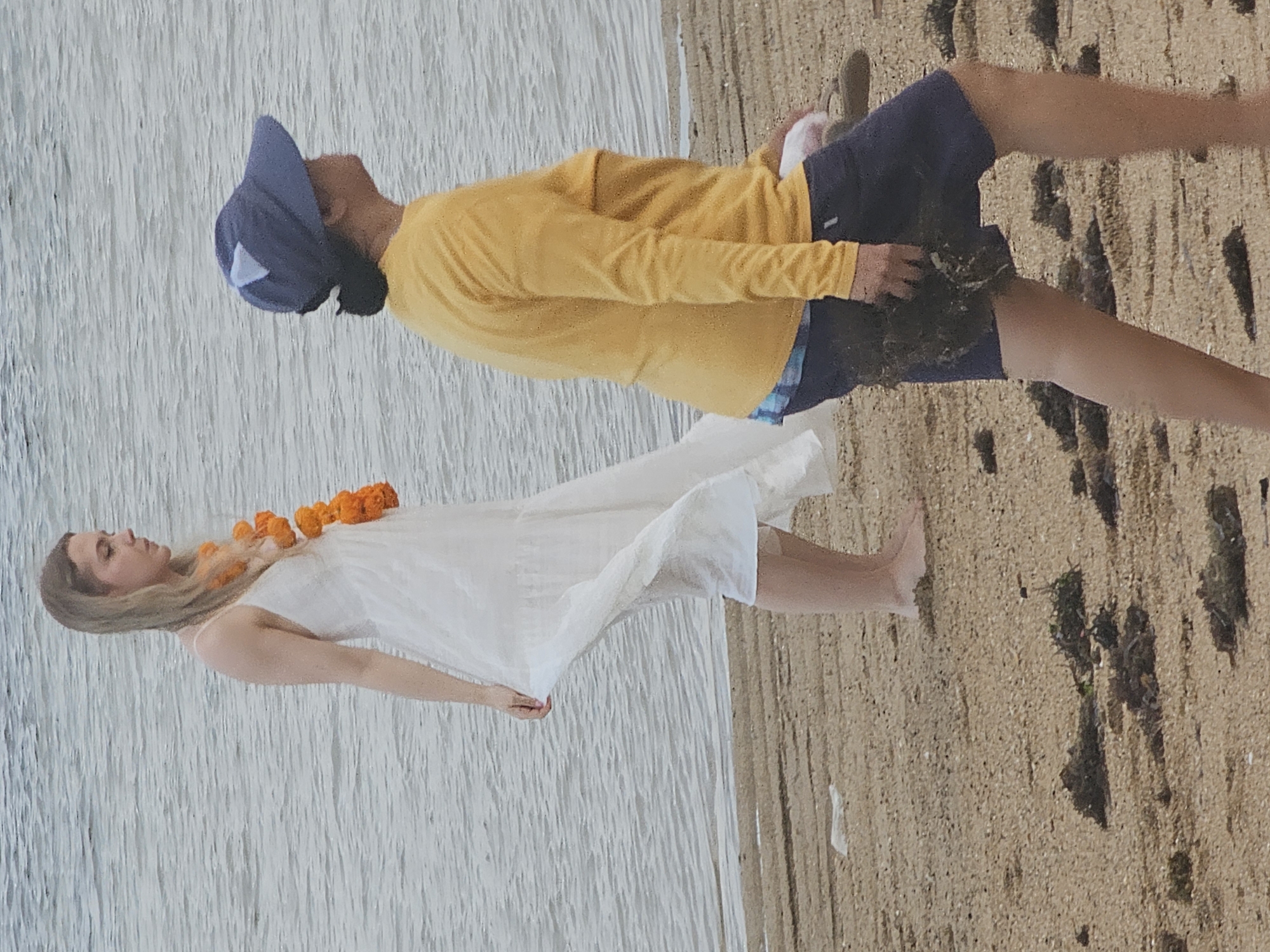 Two individuals are walking on a sandy beach near the water. On the left, a woman with blonde hair wearing a white dress with spaghetti straps and an orange flower garland walks barefoot. On the right, a person wearing a yellow long-sleeved shirt, a blue baseball cap, and dark shorts walks, holding a bundle of dark seaweed in their hand. The background is dominated by the rippling surface of the water. The beach is sandy and scattered with pieces of seaweed and debris. The overall scene suggests a casual outdoor activity. This location is likely on the coast of Denpasar, Indonesia, known for its beaches.