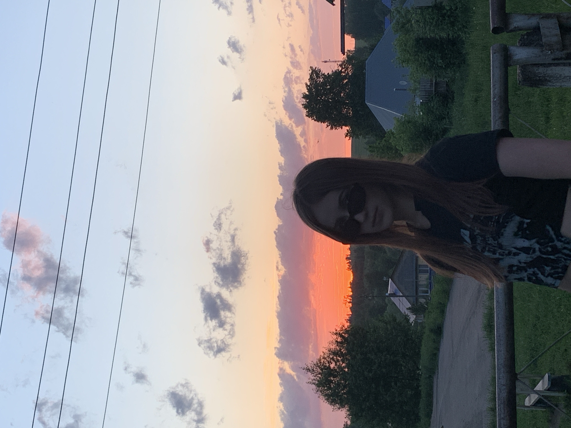 The image, taken outdoors, shows a fair-skinned person with long brown hair and sunglasses, partially visible in the right foreground. The background features a sunset sky transitioning from light blue to orange with scattered clouds. Below the sky, there is a treeline and glimpses of buildings, including a structure with a grey roof. A road or path is visible on the right side of the image. Power lines are present in the left side of the sky. The photo appears to have been taken in Фоки, Russia, based on the prompt's request.
